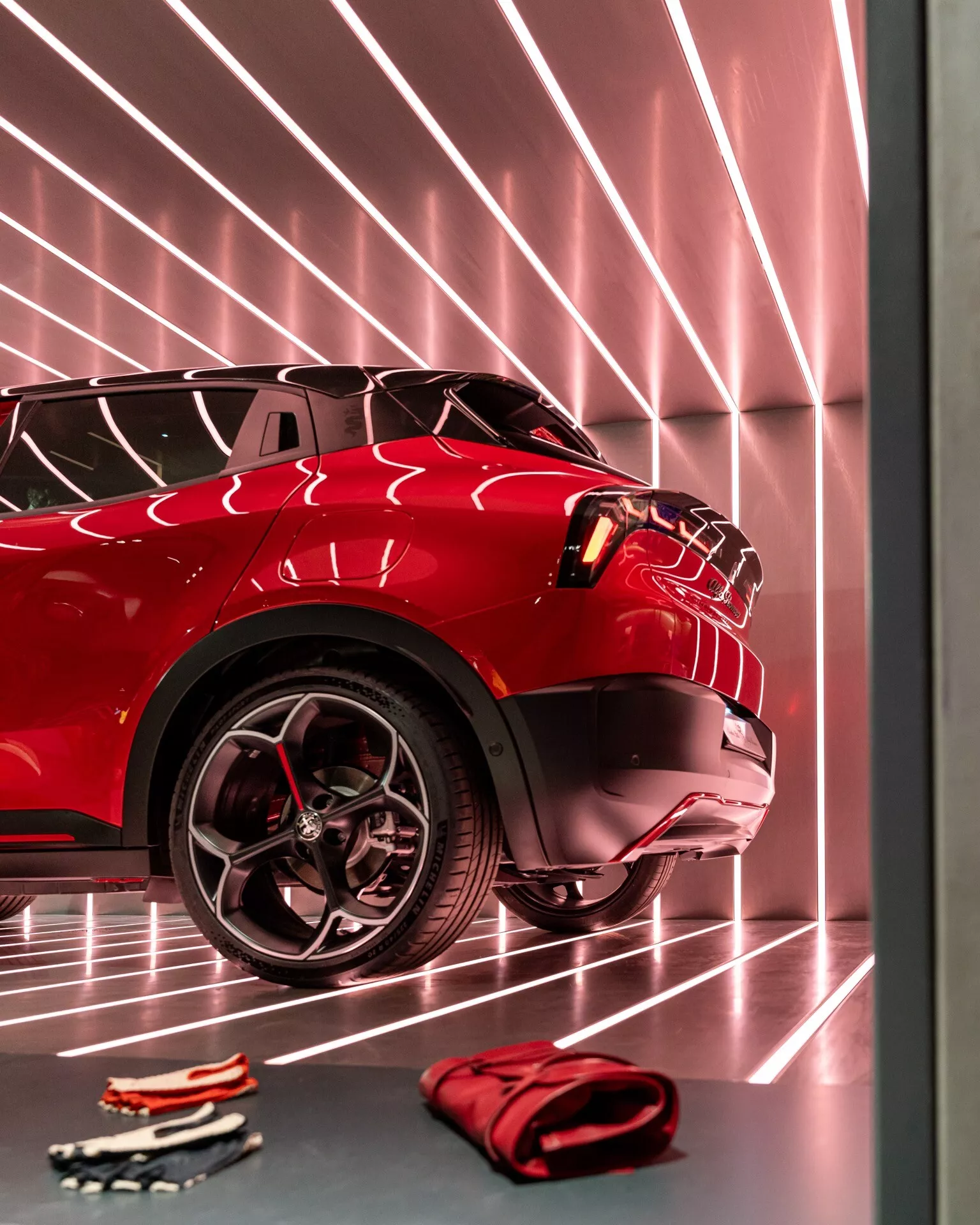 Detailní pohled na přední část červeného Alfa Romeo Junior 2024 s důrazem na pokročilý systém světlometů, sportovní design kola a odrazivý lak karoserie v interiéru showroomu