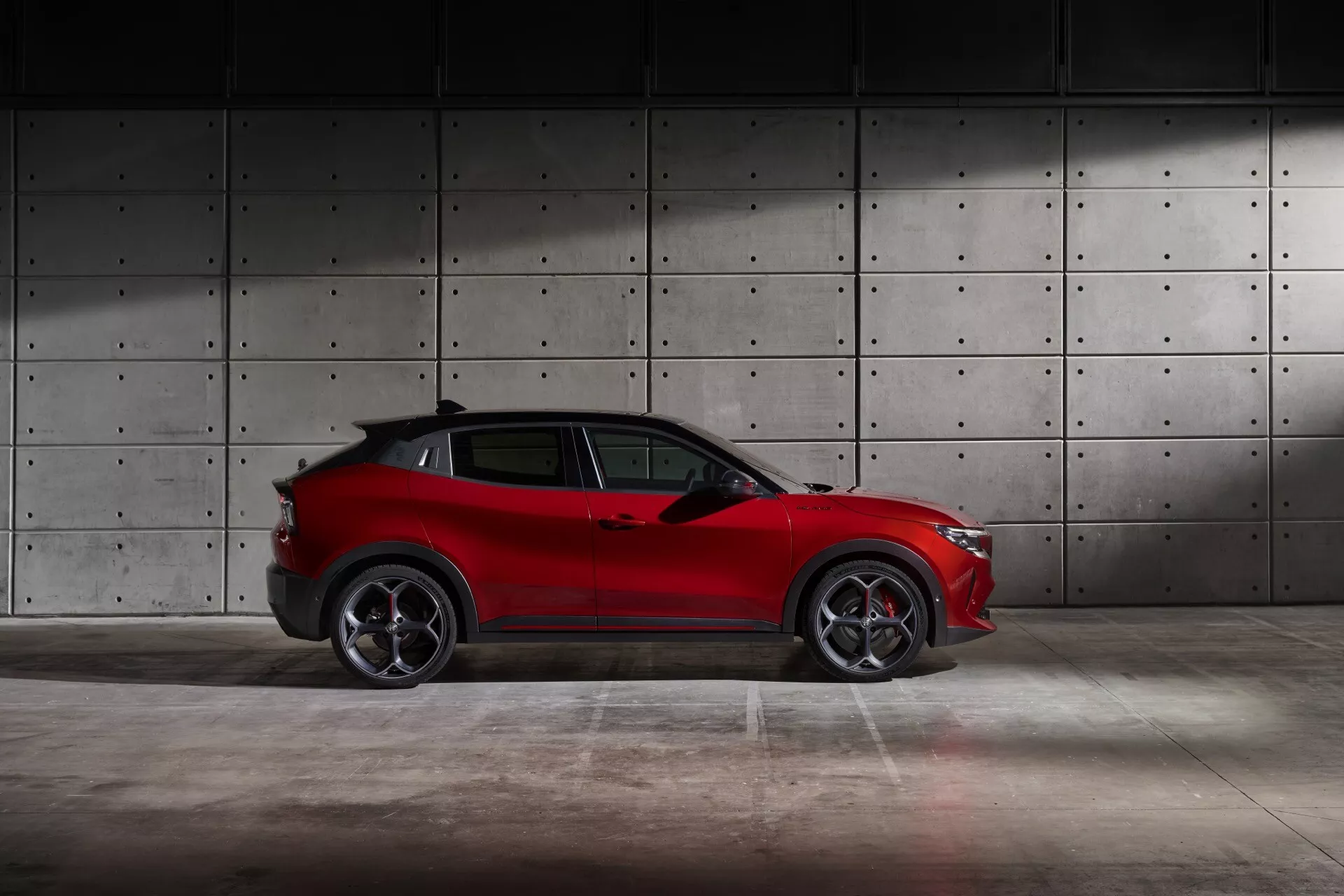 Alfa Romeo Junior 2024 v červené barvě, zobrazení z profilu zdůrazňující sportovní design SUV, sklony střechy, slitinová kola a elegantní linie karoserie na pozadí neutrální betonové stěny