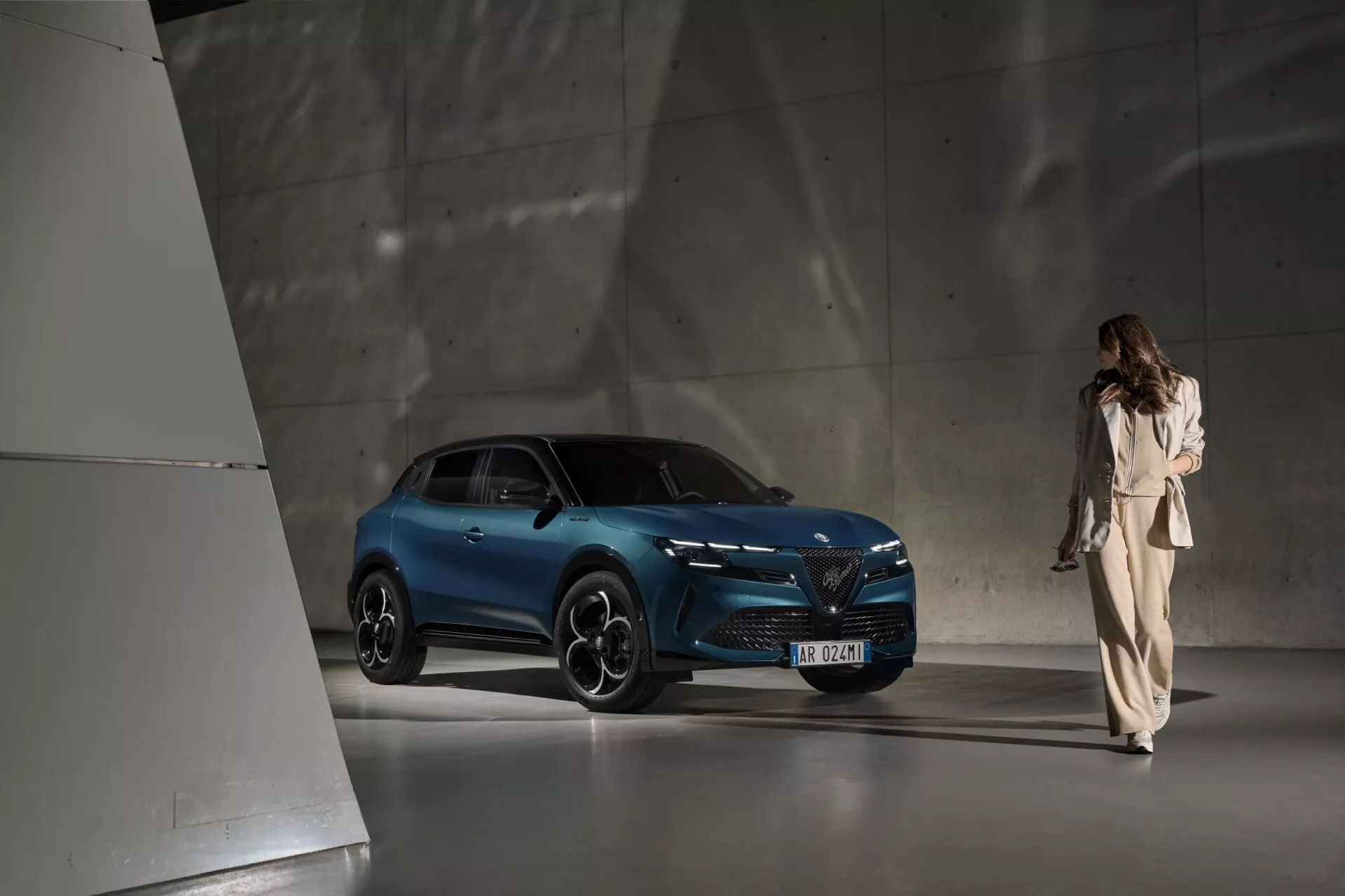 Elegantní SUV Alfa Romeo Junior 2024 představené v moderním minimalistickém showroomu s dramatičtím nadsvícením zdůrazňujícím jeho živou modrou barvu, pokročilé LED světlomety a ikonický design přední masky, zachyceno z perspektivy tří čtvrtin zepředu