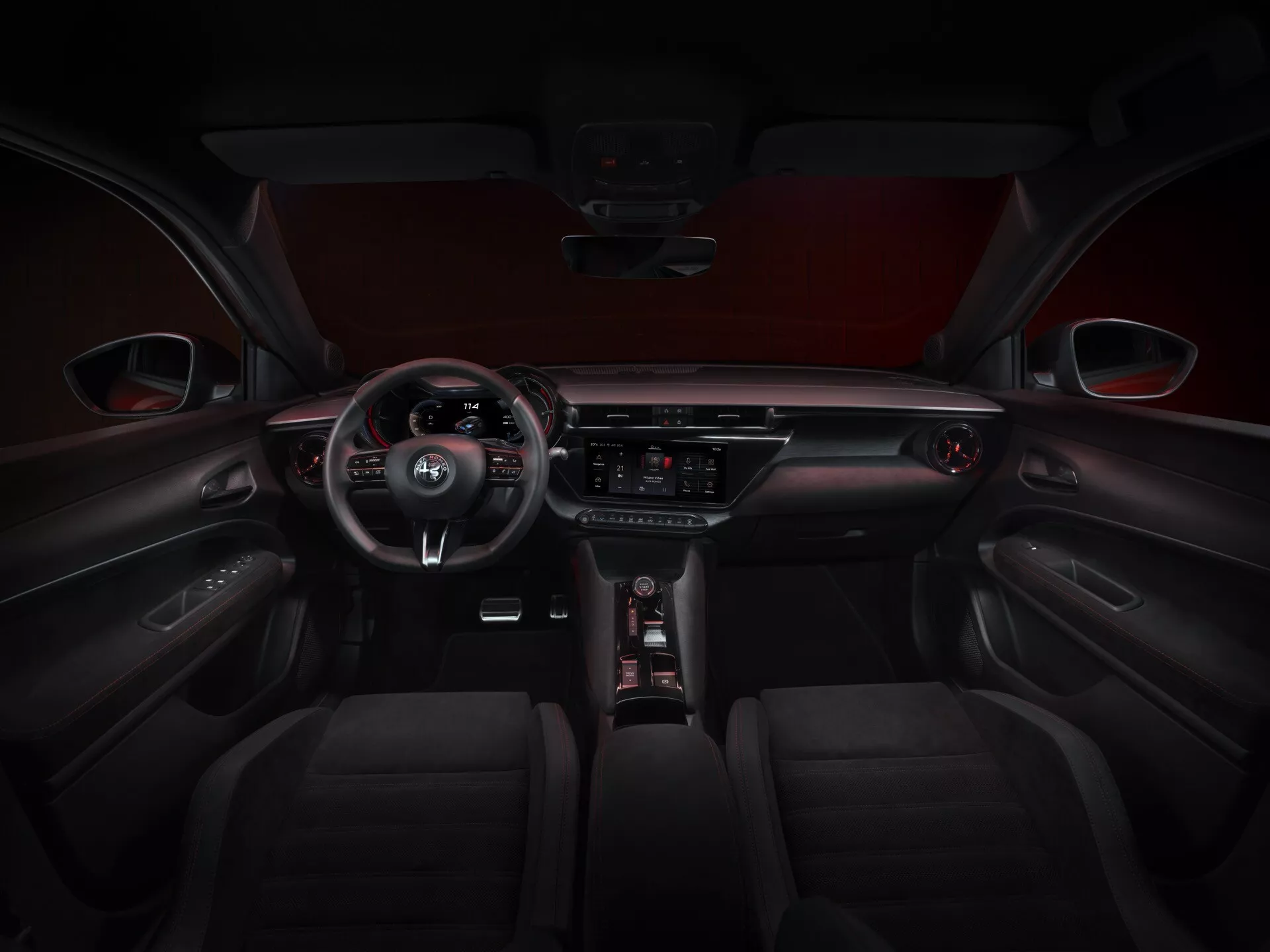 Interiérový pohled na Alfa-Romeo Junior 2024 (966) ukazující tmavý kožený potah s detaily šití na předních sedadlech, trojramenný volant s ovládáním na volantu obsahující emblem výrobce, částečně viditelný digitální přístrojový panel, centrální dotykový informačně zábavní systém s tlačítky pro ovládání klimatizace a dalších funkcí vozu pod ním, elegantní řadicí páka vedle intuitivního ovládacího kolečka, zvýrazněné kontrastním červeným čalouněním stropu a dveřními panely s integrovanými ovládáními a detaily šití.