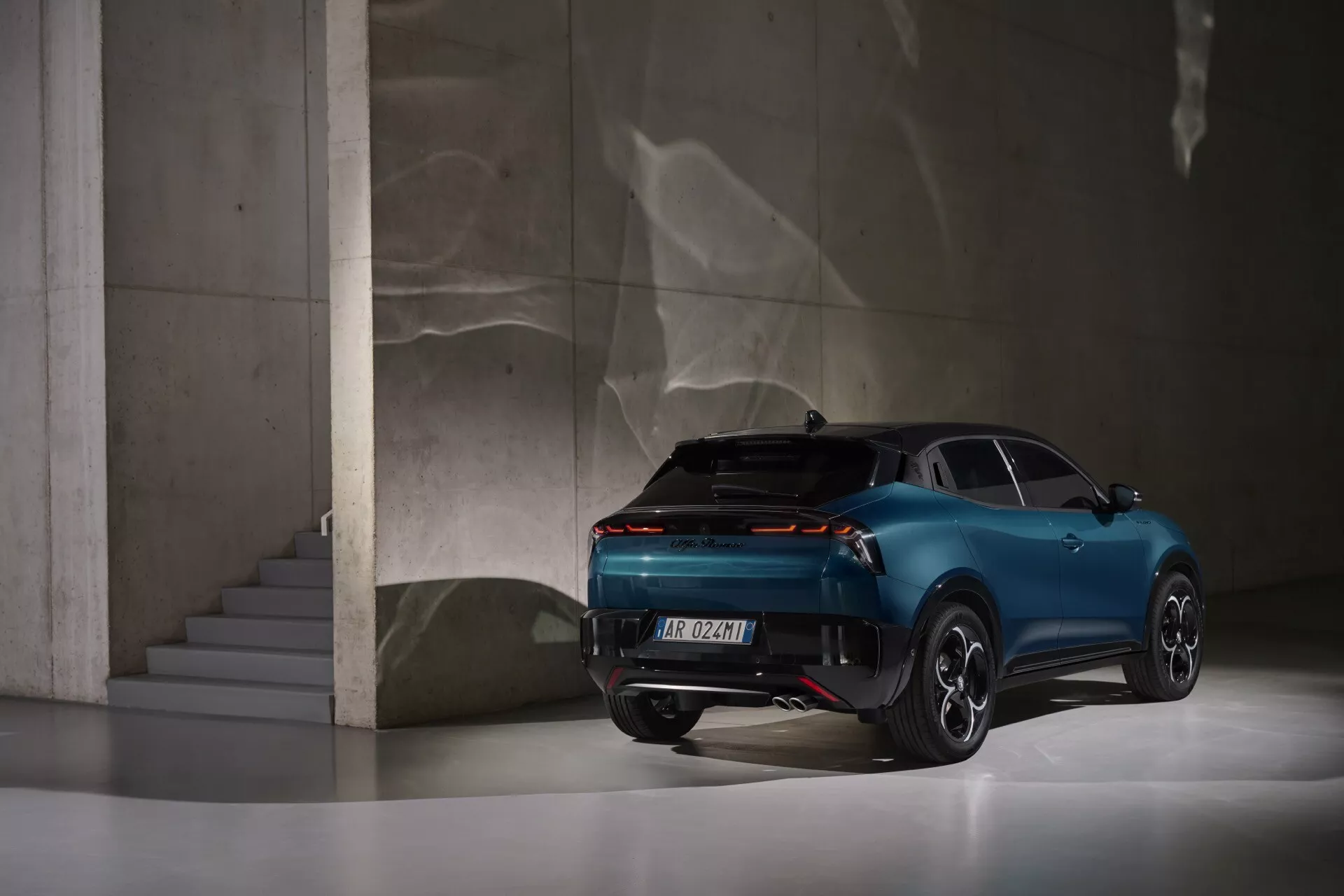 Alfa Romeo Junior 2024 zobrazený ze zadního tříčtvrtečního pohledu, který ukazuje jeho moderní design, zadní světla pokrývající téměř celou šířku vozu, sportovně-kupéový siluetu s čtyřmi dveřmi a luxusní prvky, umístěný v elegantním interiéru s odrazivou podlahou a betonovými konstrukcemi.