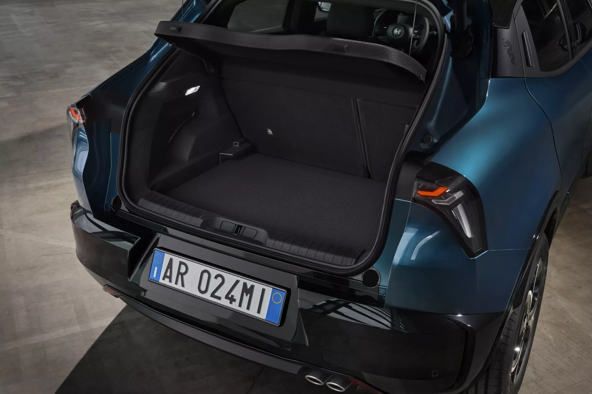 Zadní pohled na Alfa Romeo Junior 2024 ukazující prázdný kufr, metalickou modrou barvu, moderní design s aerodynamickými liniemi, integrovaná brzdová světla na zadním víku, LED zadní světla a dvojitý výfukový systém.