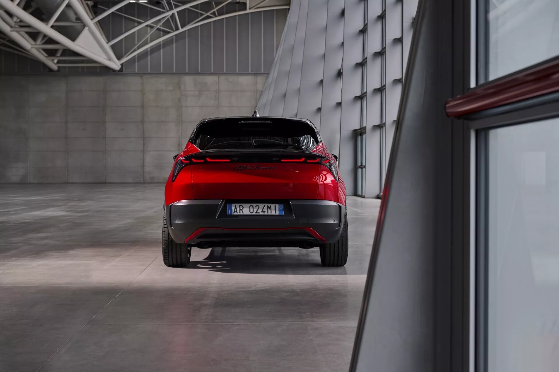 Zadní pohled na Alfa Romeo Junior 2024 zobrazující designové prvky a LED zadní světla v průmyslovém prostředí s leštěnou podlahou a ambientním osvětlením