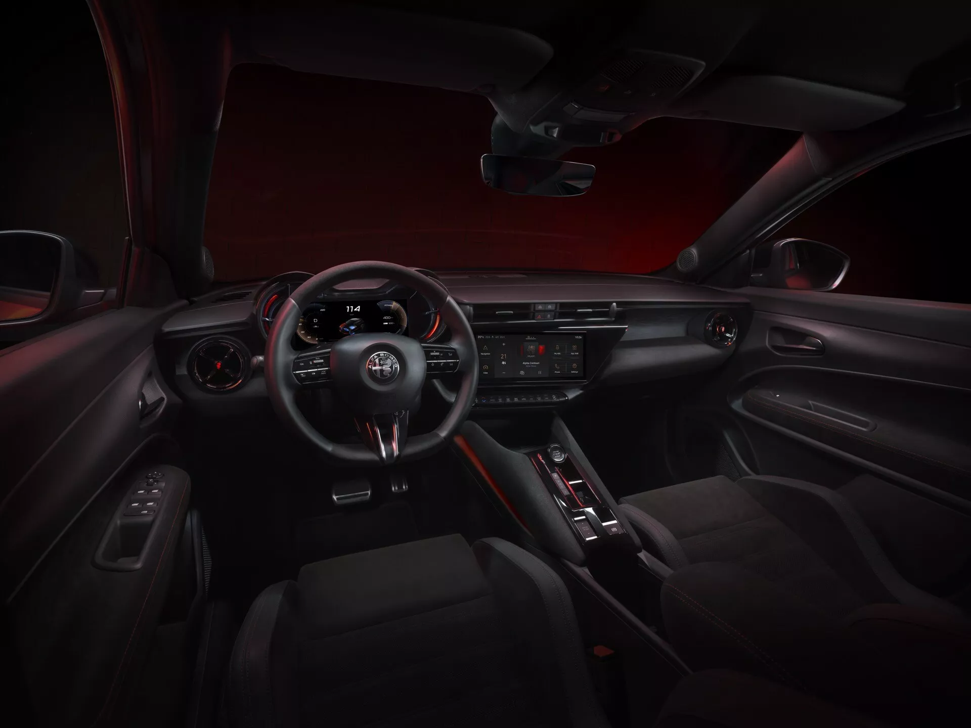 Interiérový pohled na kokpit Alfa-Romeo Junior 2024 z centrálního zadního sedadla, zobrazující palubní desku, sportovní volant se třemi rameny s integrovanými ovladači a pádly pro řazení, přístrojový panel, centrální displej informačního systému, tlačítka klimatizace, volič automatické převodovky, dveřní panel s ovládáním a sportovně elegantní atmosféru s červeným ambientním osvětlením