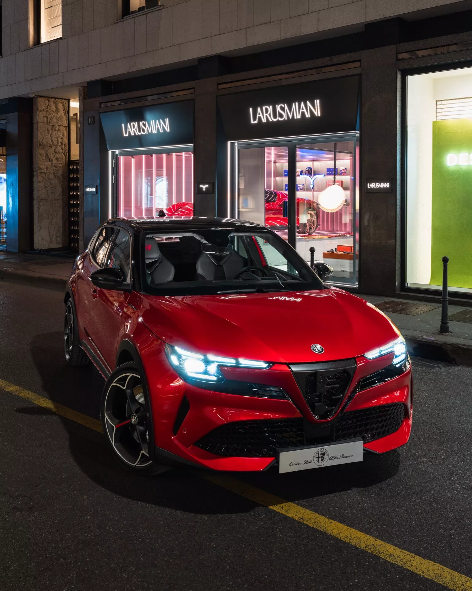 Červené sportovní užitkové vozidlo Alfa-Romeo Junior 2024 zaparkované na městské ulici v noci, předvádějící jeho sportovní a agresivní design s výraznou mřížkou, moderními LED světlomety a koly z lehkých slitin orientovanými na výkon