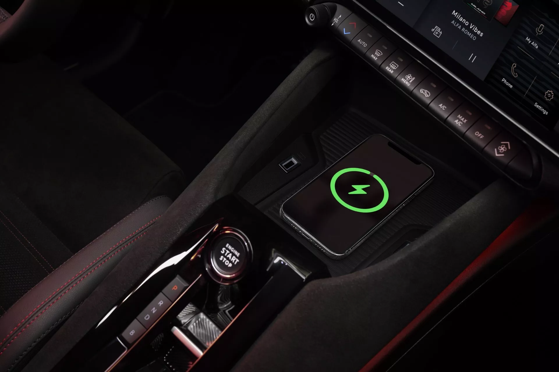 Pohled shora na středovou konzolu Alfa-Romeo Junior 2024 s červeným prošíváním, elegantní volič rychlostí s indikacemi automatické převodovky, bezdrátovou nabíjecí podložkou se zeleným symbolem blesku, tlačítky ovládání, ambientním osvětlením, držákem na nápoje s červenými akcenty, což zdůrazňuje luxusní a technologicky orientovaný design interiéru