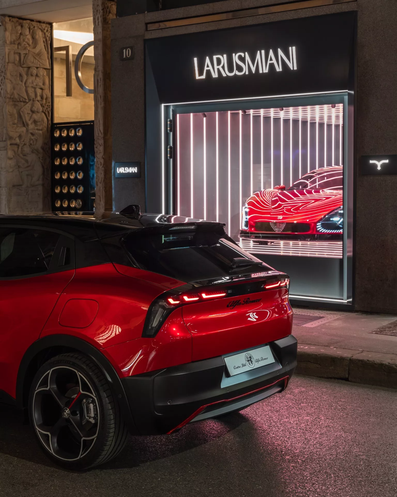 Zadní pohled z tříčtvrteční perspektivy na moderní červený Alfa Romeo Junior 2024 zaparkovaný v noci před obchodem Larusmani, zobrazující LED zadní světla, zadní čelní sklo se stěračem, střešní spoiler, slitinová kola s nízkoprofilovými pneumatikami a naznačující technologii elektrického nebo hybridního vozidla, ve městském osvětlení.