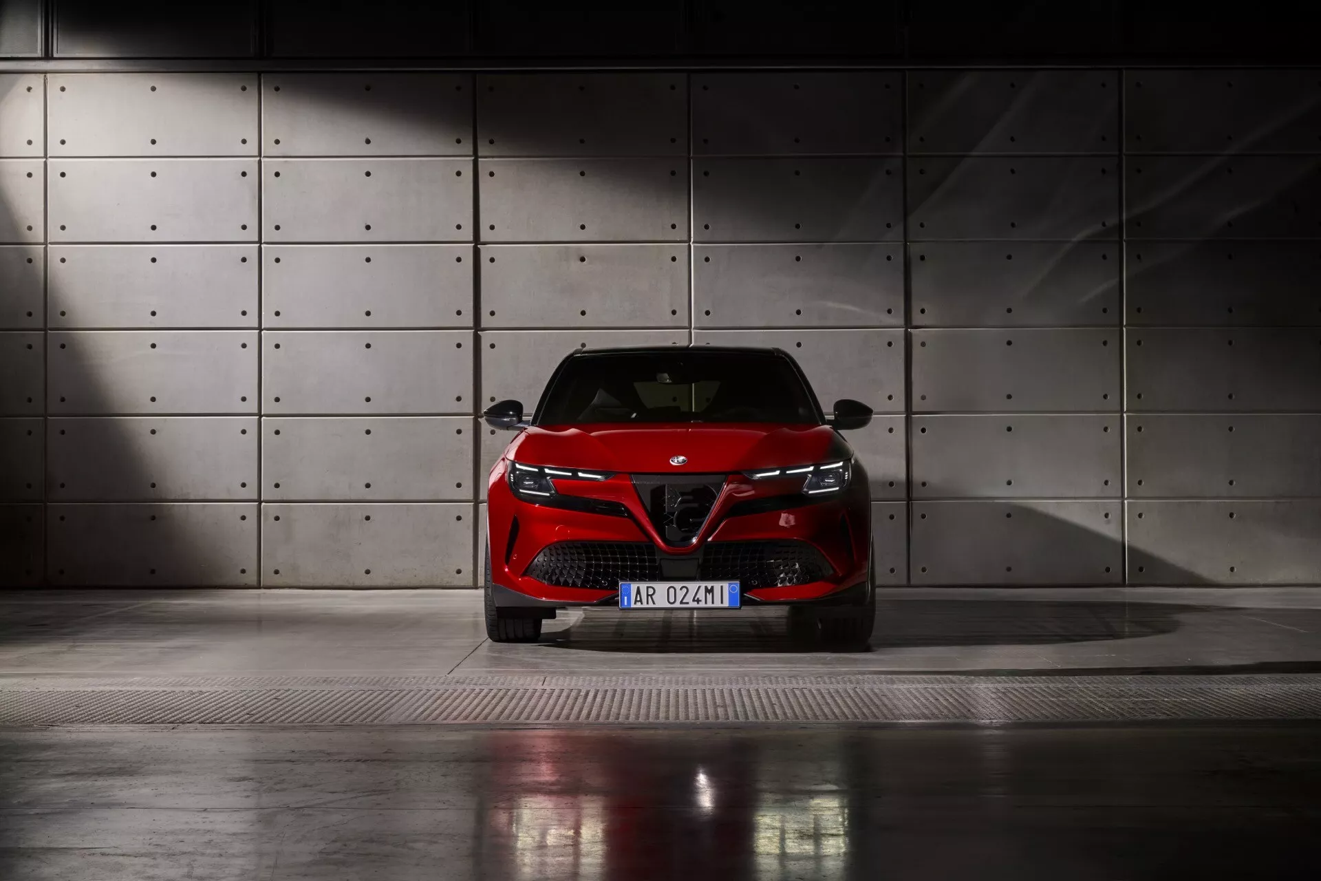 Pohled zepředu na Alfa Romeo Junior 2024 zachycený z nízkého úhlu na kovové mřížkové podlaze, s LED světlomety, výraznou mřížkou chladiče se znakem výrobce a elegantním designem nárazníku, na pozadí betonové stěny s kruhovými prohlubněmi pod dramatickým osvětlením.