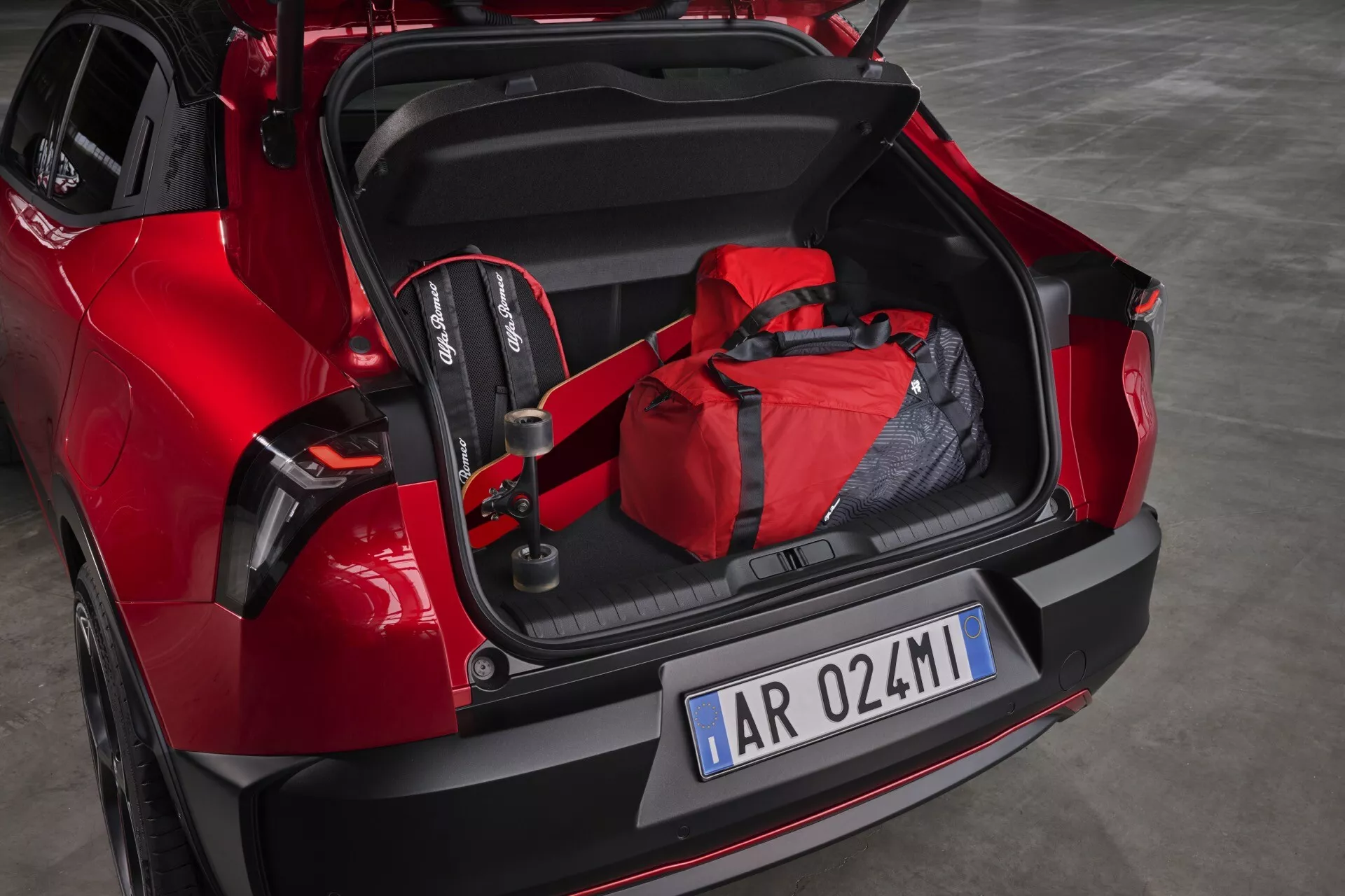 Model Alfa-Romeo Junior 2024 v červené barvě s otevřeným kufrem ukazujícím prostorný interiér, dětskou autosedačku a červenou sportovní tašku, se sportovním zadním designem a moderními LED zadními světly, což demonstruje potenciál pro rodinnou přívětivost a technologii asistence při parkování