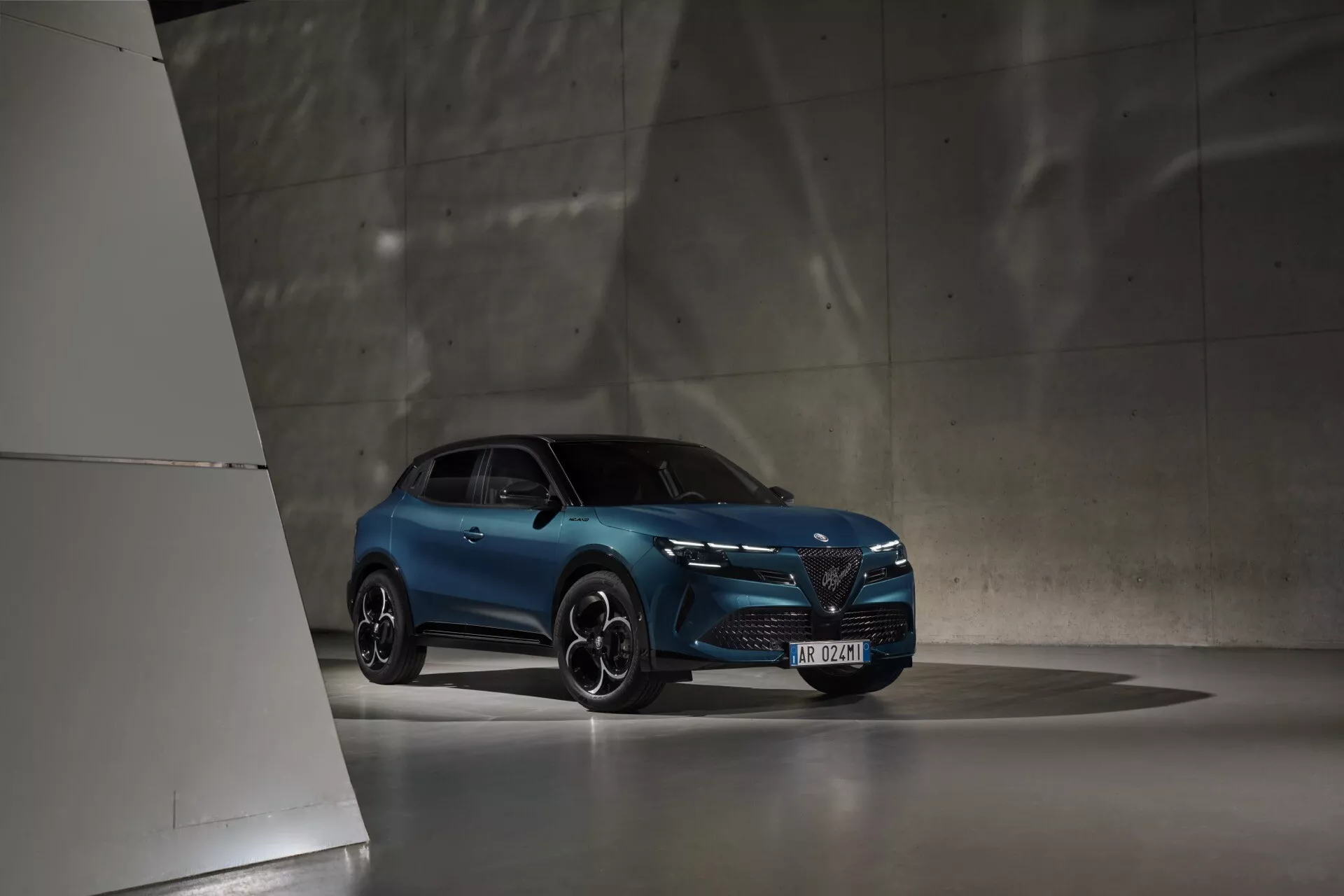 Modrý Alfa Romeo Junior z roku 2024 v špatně nasvíceném interiéru, zachycený z nízkého úhlu zdůrazňující jeho moderní design a sportovní postoj, s LED světlomety, výraznou přední mřížkou a sportovními slitinovými koly.