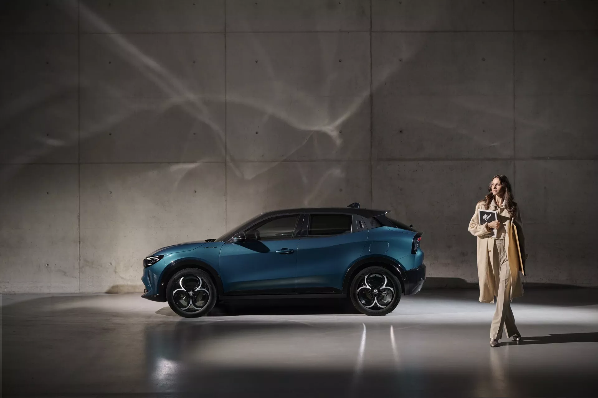 Dynamický boční pohled na Alfa Romeo Junior 966 z roku 2024 v metalické modré, zdůrazňující jeho elegantní design a pokročilé funkce, včetně sportovních slitinových kol a aerodynamického profilu, v sofistikovaném vnitřním prostředí s dramatickým osvětlením