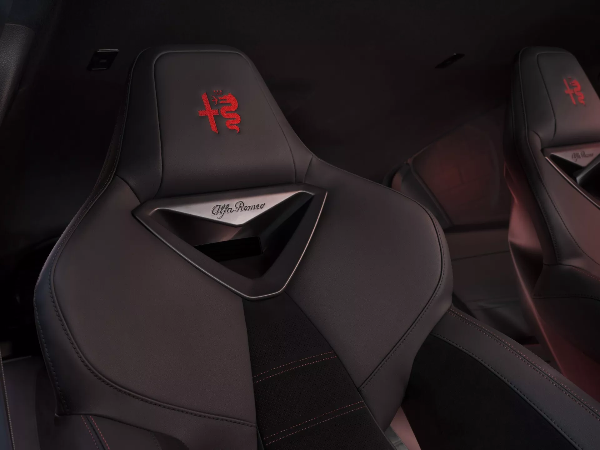 Detailní pohled na černá sportovní sedadla Alfa Romeo Junior 2024 s vyšívaným červeným logem '+' na opěrce hlavy a červeným kontrastním prošíváním, ukazující luxusní a sportovní design interiéru
