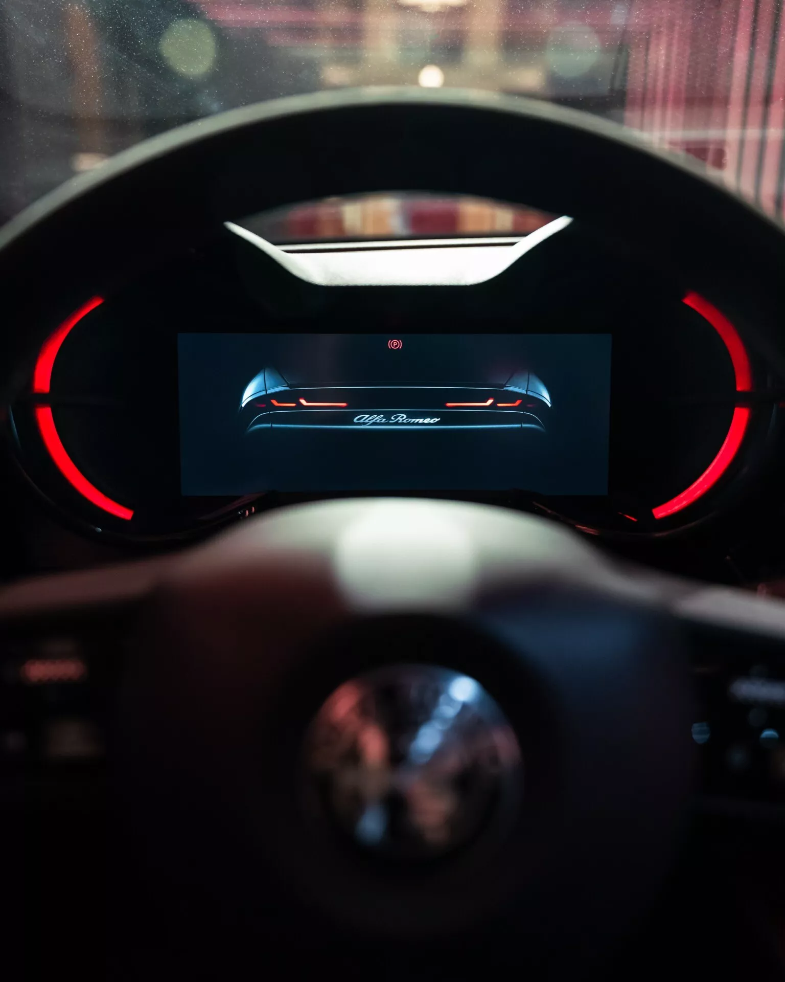 Interiérový pohled na Alfa-Romeo Junior 2024 se zaměřením na digitální palubní desku vysokého rozlišení řidiče s červenými akcenty a částečným pohledem na volant s rozmazaným pozadím naznačujícím luxusní prostředí