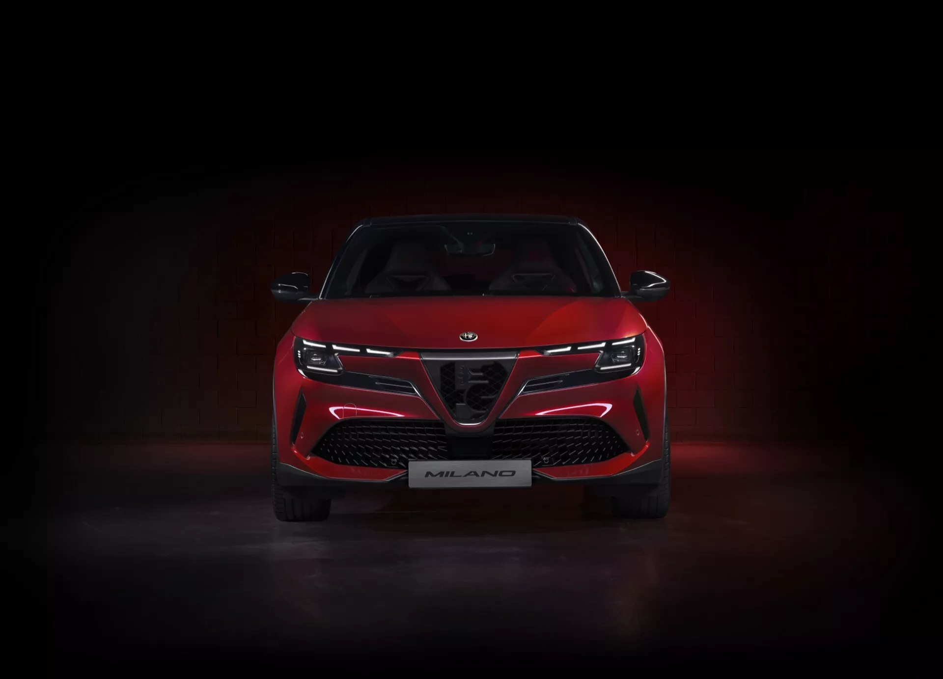 Přední pohled na červený Alfa-Romeo Junior 2024 (966), osvětlený ve studiu na tmavém pozadí s leštěným podsvícením, který předvádí jeho charakteristickou mřížku s velkými vzduchovými vstupy, úzké elegantní LED světlomety propojené světelným pásem a odznak vydání Milano, což zdůrazňuje jeho sportovní a agresivní designové prvky.