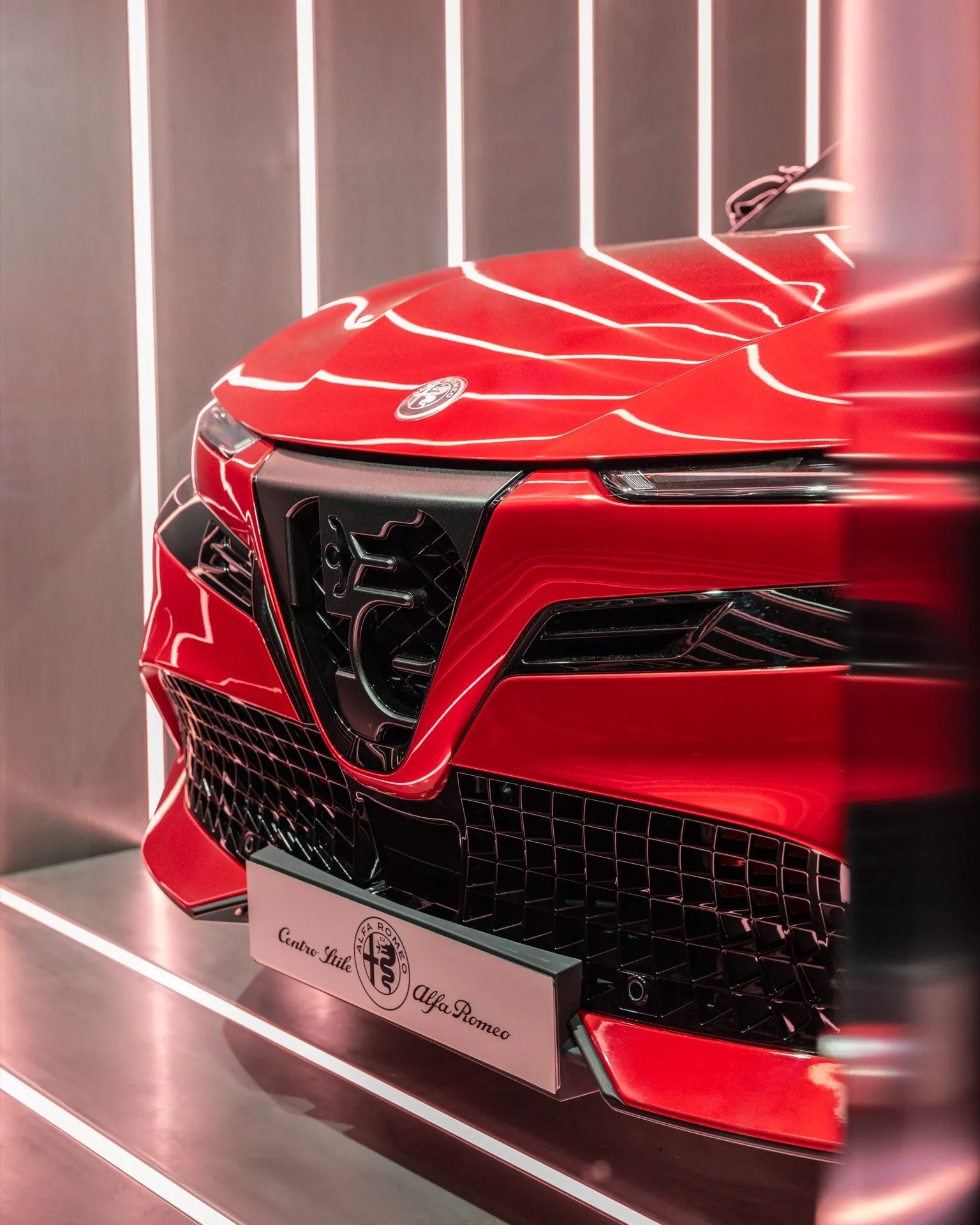 Detailní obrázek přední levé části Alfa-Romeo Junior 2024 s živou metalickou červenou barvou, zobrazující jeho agresivní design, ostré úhly, tvarovaný povrch, výrazný emblém, velkou úhlovou mřížku, elegantní denní svícení a možné výkonnostní sání vzduchu, na pozadí jednoduchého interiéru pravděpodobně showroomu.