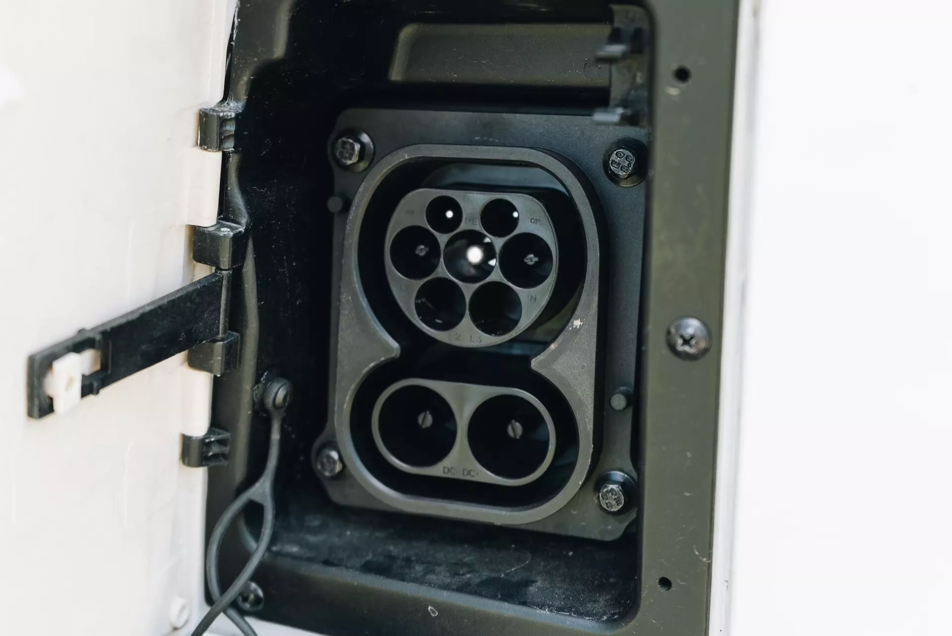 Detailní pohled na zásuvku nabíjecího konektoru elektromobilu (EV) zobrazující dva typy konektorů: konektor typu 2 se sedmipinovým uspořádáním a konektor CHAdeMO se čtyřmi velkými piny a menším komunikačním pinem, což naznačuje kompatibilitu s více standardy nabíjení EV. Zásuvka má otevřené zavěšené víko a připojen flexibilní kabel, zdůrazňující zaměření na infrastrukturu EV, technologii nabíjení a udržitelnou dopravu.