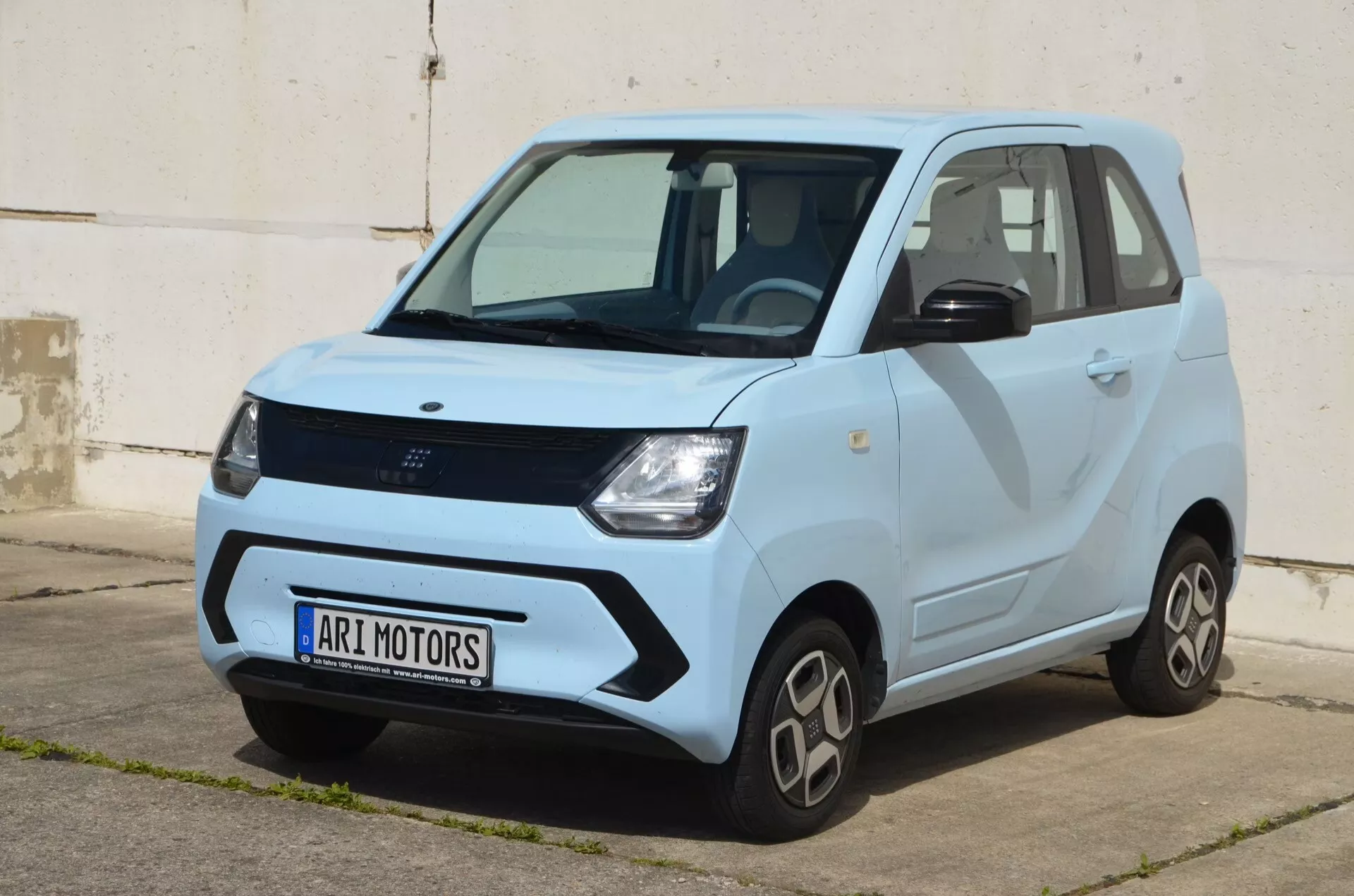 Exteriérový tříčtvrteční přední pohled na Ari-Motors Soleno 2024, který ukazuje jeho kompaktní elektrický design. Obraz zdůrazňuje oblast čelní masky, světlomety, přední kolo a profil řidiče v přirozeném světle, což odhaluje linie karoserie a kontrastní barevné schéma s tmavším tónem na spodní polovině. Jsou viditelné praktické designové prvky, jako jsou rozšířené oblouky kol, kola skromné velikosti a výrazné kliky dveří. Zřetelný záhyb běží od předního oblouku kola ke zadní části, což dodává charakter. Malá velikost naznačuje vhodnost pro městské prostředí.