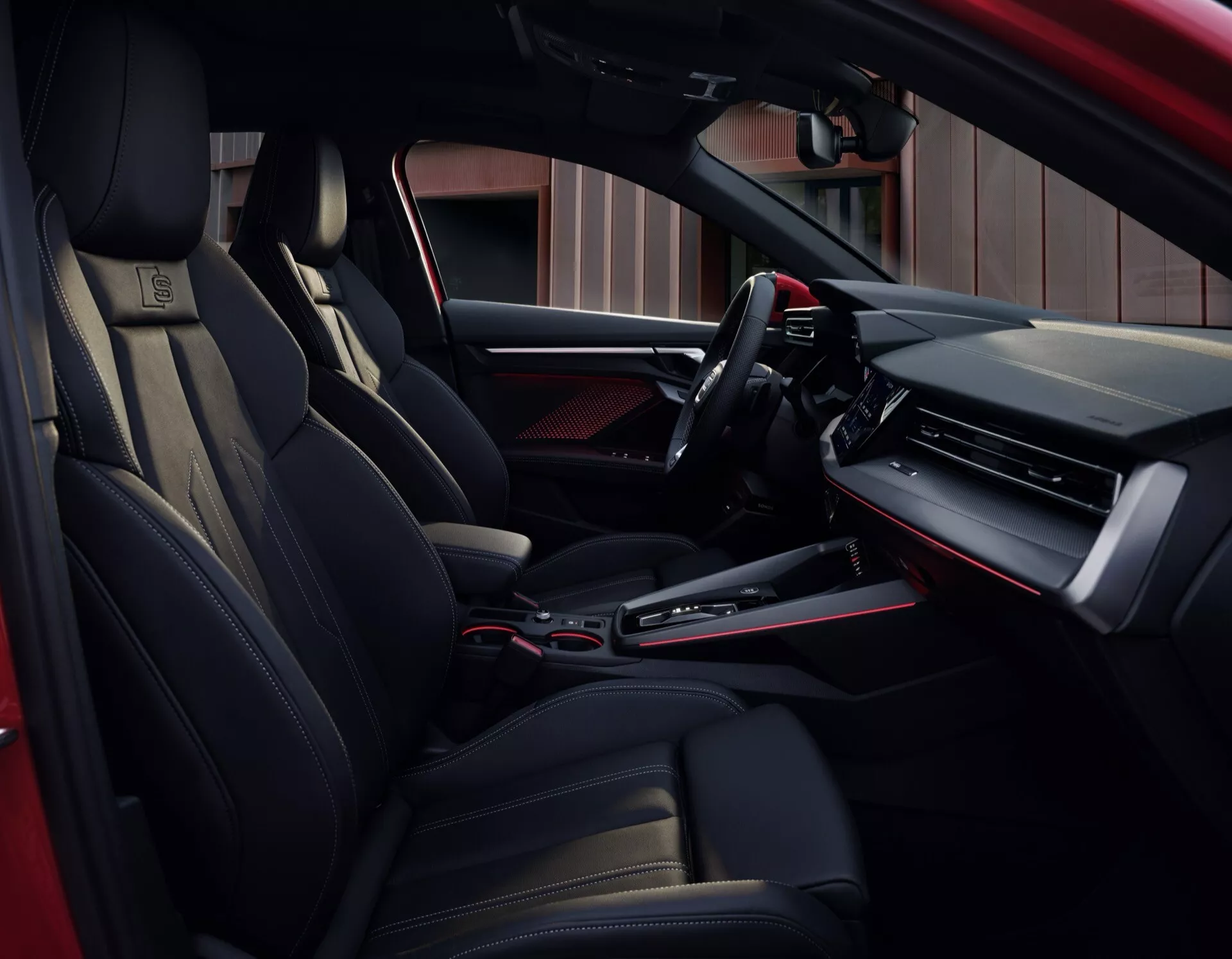 Interiérový pohled na Audi A3 2024 (8Y-facelift) zaměřený na stranu řidiče z pohledu zadního cestujícího, zobrazující elegantní palubní desku s digitálním displejem a velkou obrazovkou infotainmentu, multifunkční volant s pokročilými ovládacími prvky, sportovní ergonomická sedadla s kůží, automatická převodovka v centrální konzole a luxusní černé a kovové akcenty po celé kabině.