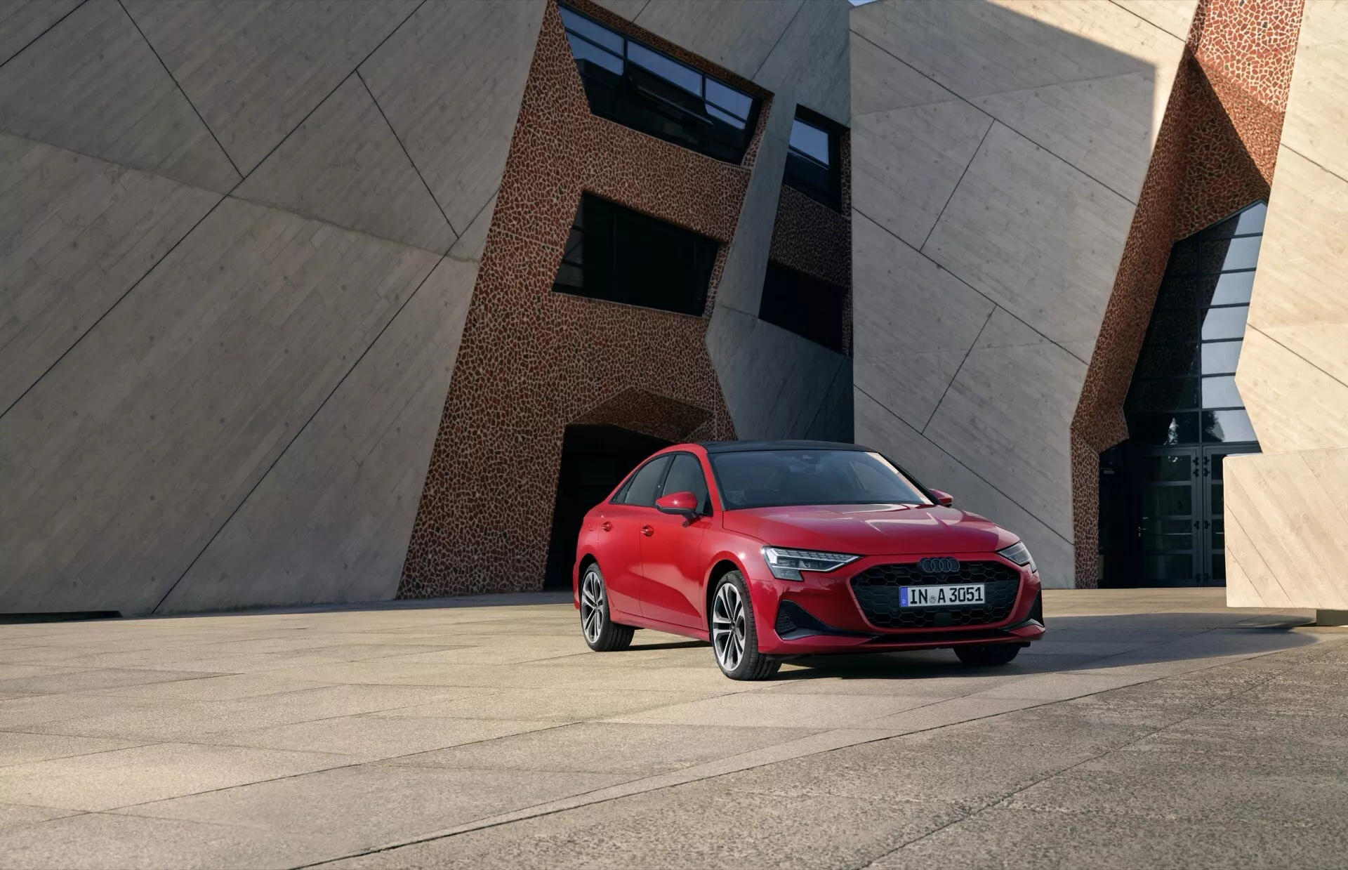 Audi A3 8Y Facelift 2024 v jasně červené barvě, předvádějící LED světlomety, výraznou masku chladiče a slitinová kola na pozadí moderního městského prostranství a geometrické architektury