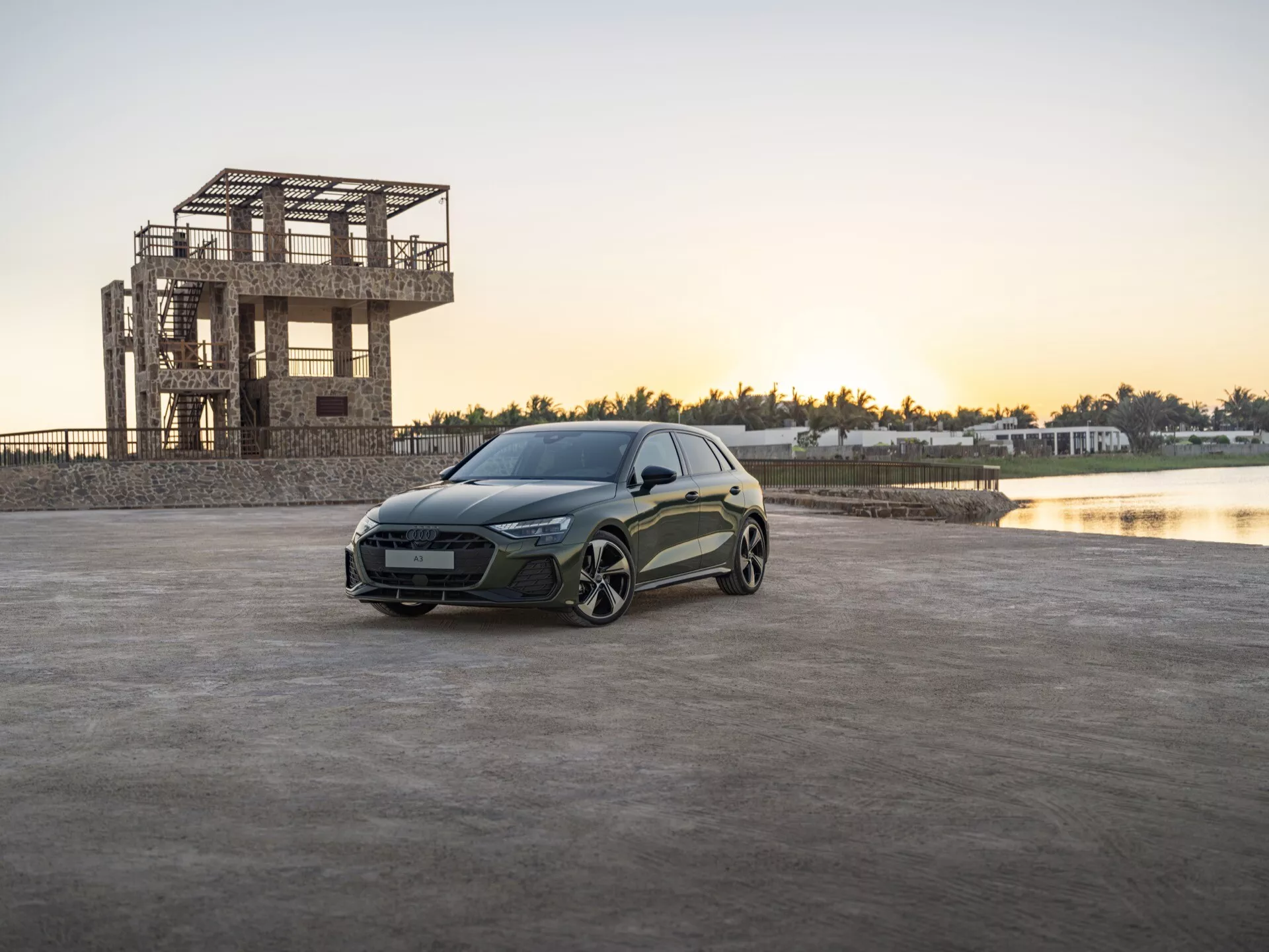 Audi A3 (8Y-Facelift) 2024 umístěné na otevřené zpevněné ploše za svítání nebo za soumraku s budovou ve výstavbě a vodní plohou na pozadí, předvádějící vozidlo s elegantními LED světlomety, sochařsky zpracovaným karoserií a výkonnostně orientovanými slitinovými koly.