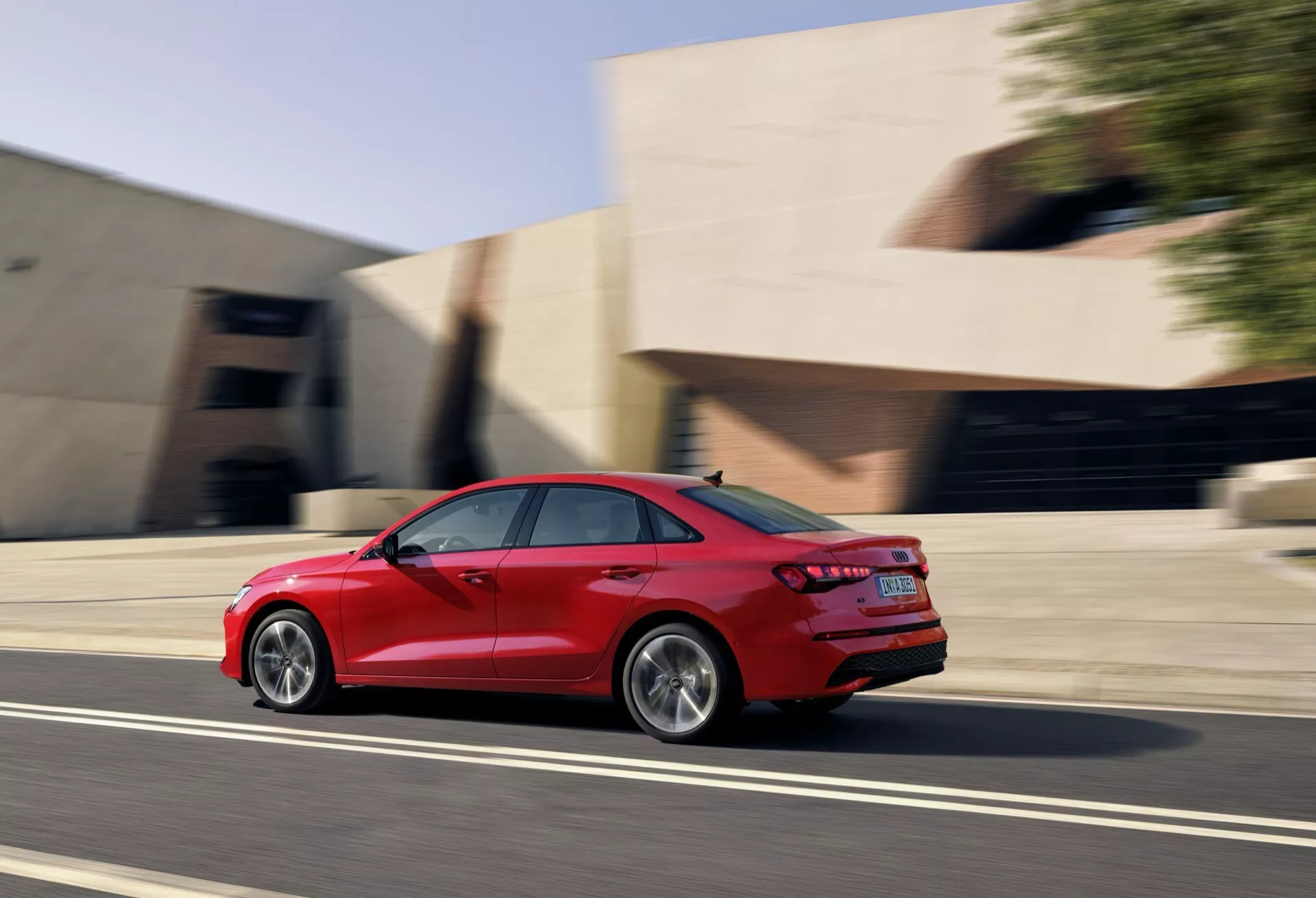 Audi A3 sedan 2024 v pohybu, červená barva, zachycený z pohledu ze tříčtvrteční zadní perspektivy ukazující slitinová kola, aerodynamický design karoserie, LED zadní světla, dvojité výfuky a efekt rozostření pohybu na městské ulici během zlaté hodiny