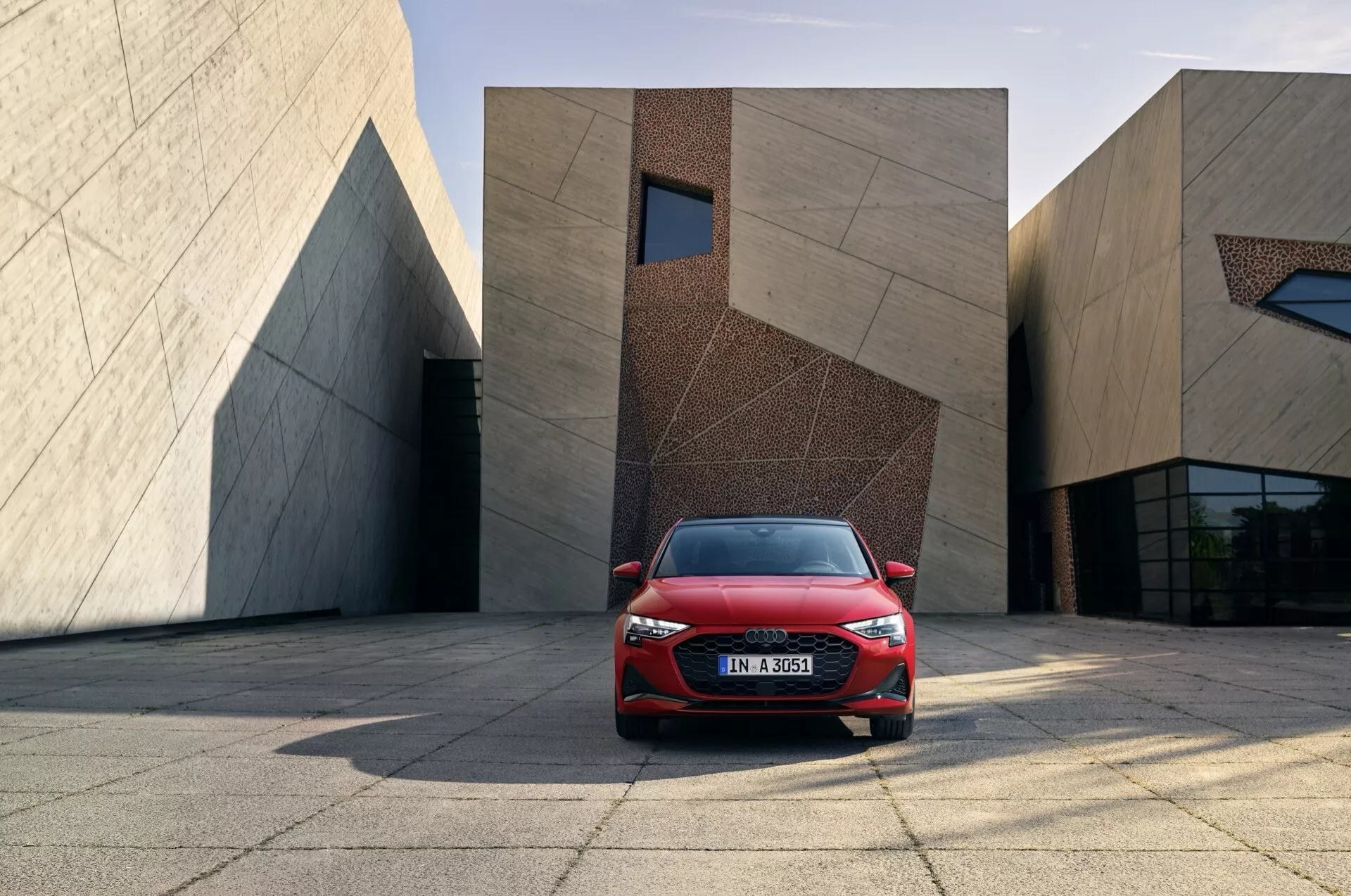 Červený Audi A3 (8Y-Facelift) rok 2024 s moderními LED světlomety a aerodynamickým designem, umístěný na městském náměstí s moderními budovami na pozadí
