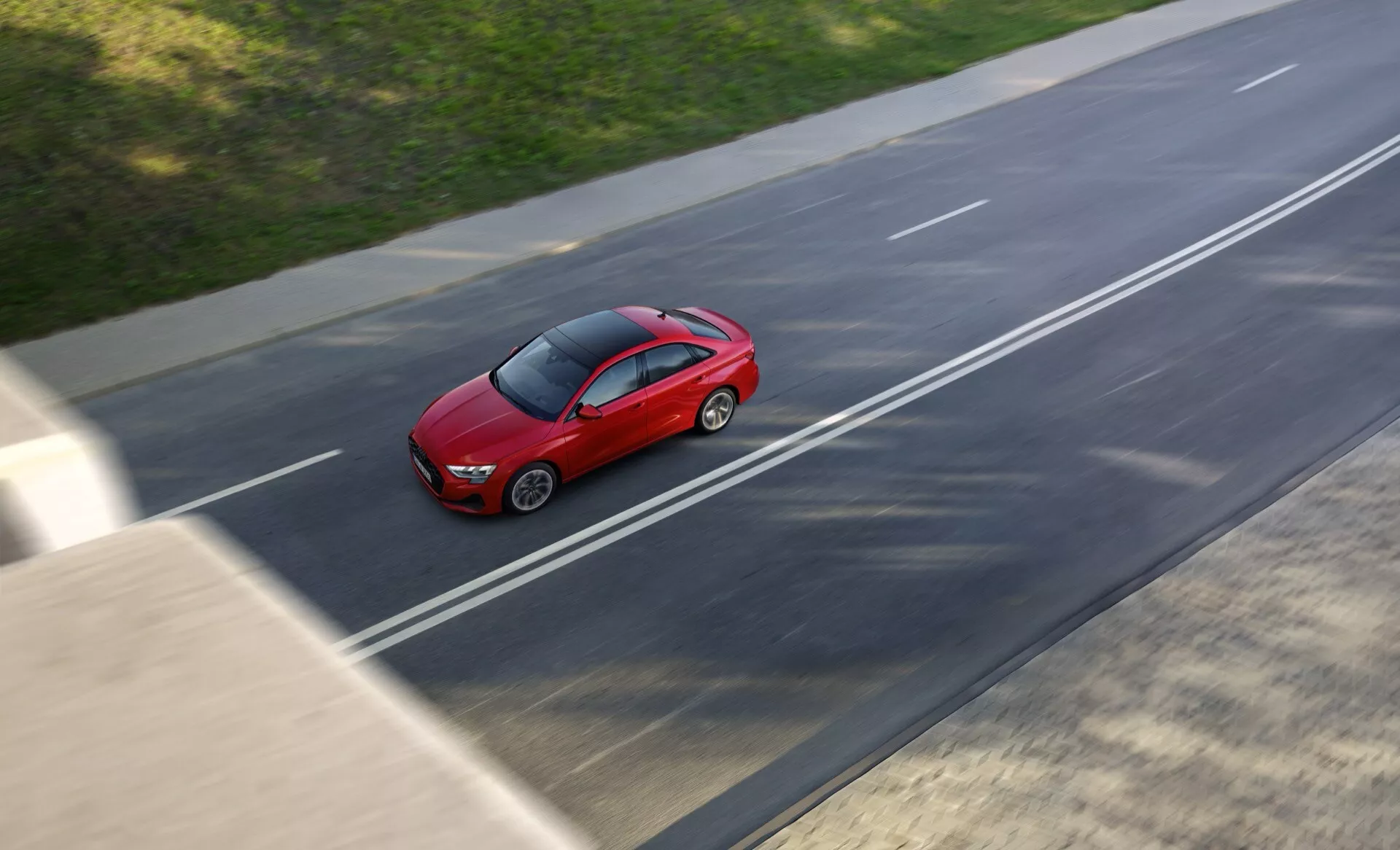 Model Audi A3 8Y facelift 2024 v jasně červené barvě, zachycený z ptačí perspektivy při jízdě po víceproudé dálnici, s mírným rozostřením pohybu a vystavující elegantní, aerodynamický design sedanu za slunečných podmínek