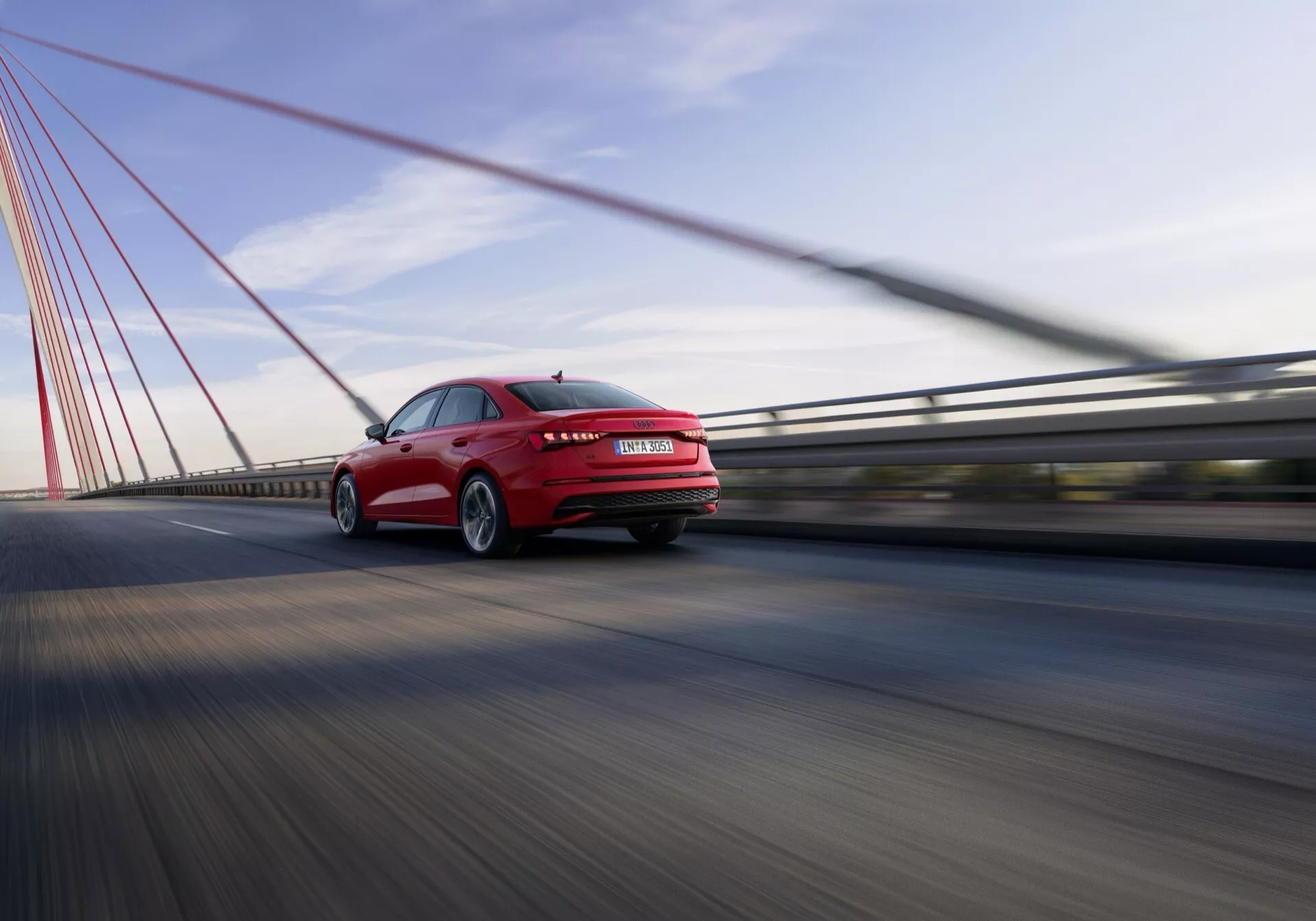 Dynamický pohled zezadu a ze strany na červený sedan Audi A3 2024 na visutém mostě, ukazuje rychlost, slitinová kola, LED zadní světla a vysoce kvalitní lak