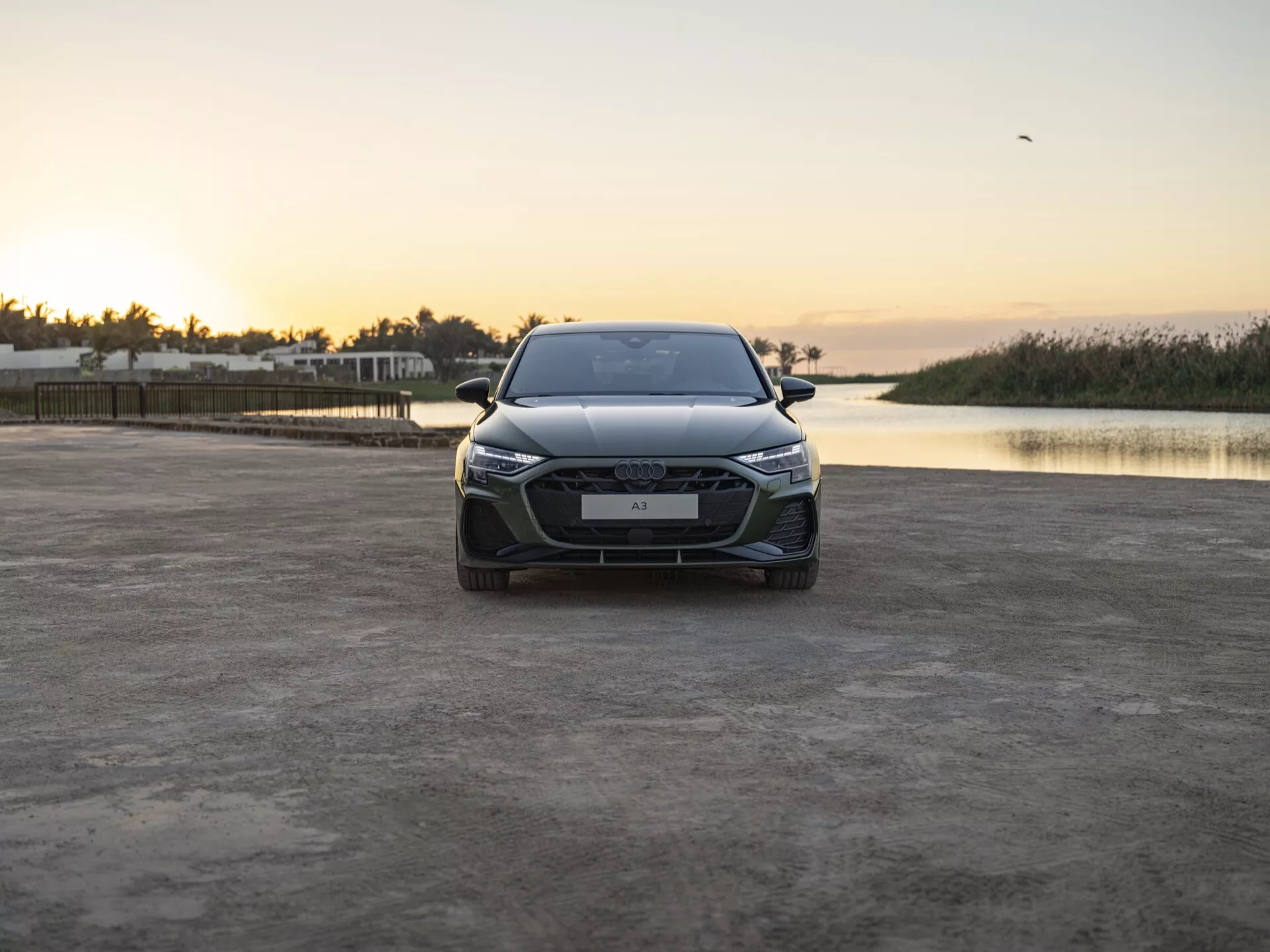 Přední pohled na Audi A3 (8Y-Facelift) rok 2024 na nezpevněném povrchu s výraznou mřížkou, LED světlomety s denními svíceními, pokročilými systémy asistence řidiče na nárazníku a tvarovanou kapotou, během klidného východu nebo západu slunce s klidnou vodní hladinou na pozadí a osamělým ptákem na obloze, což zdůrazňuje moderní design a technologické pokroky vozu proti přírodní scenerii