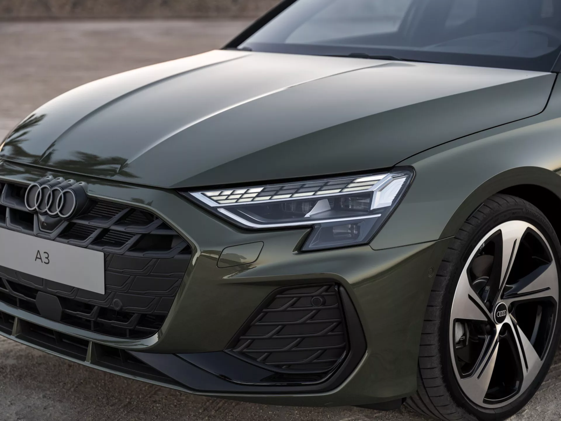 Detailní pohled na přední levou část Audi A3 2024 (8Y-Facelift) s výraznými liniemi kapoty, levým LED světlometem, šestiúhelníkovou přední mřížkou se vodorovnými lamelami a naznačeným výkonnostně orientovaným slitinovým kolem a blatníkem