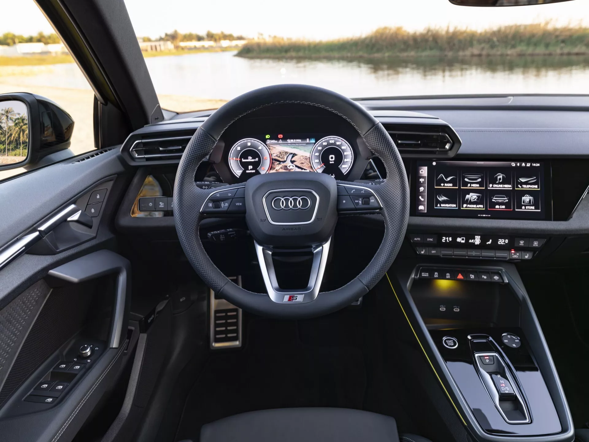Interiér Audi A3 (8Y-Facelift) 2024 z pohledu řidiče s sportovním multifunkčním volantem potaženým kůží, plně digitálním přístrojovým štítem Audi Virtual Cockpit a dotykovým infotainment systémem na palubní desce, umístěný proti klidnému venkovnímu prostředí u jezera