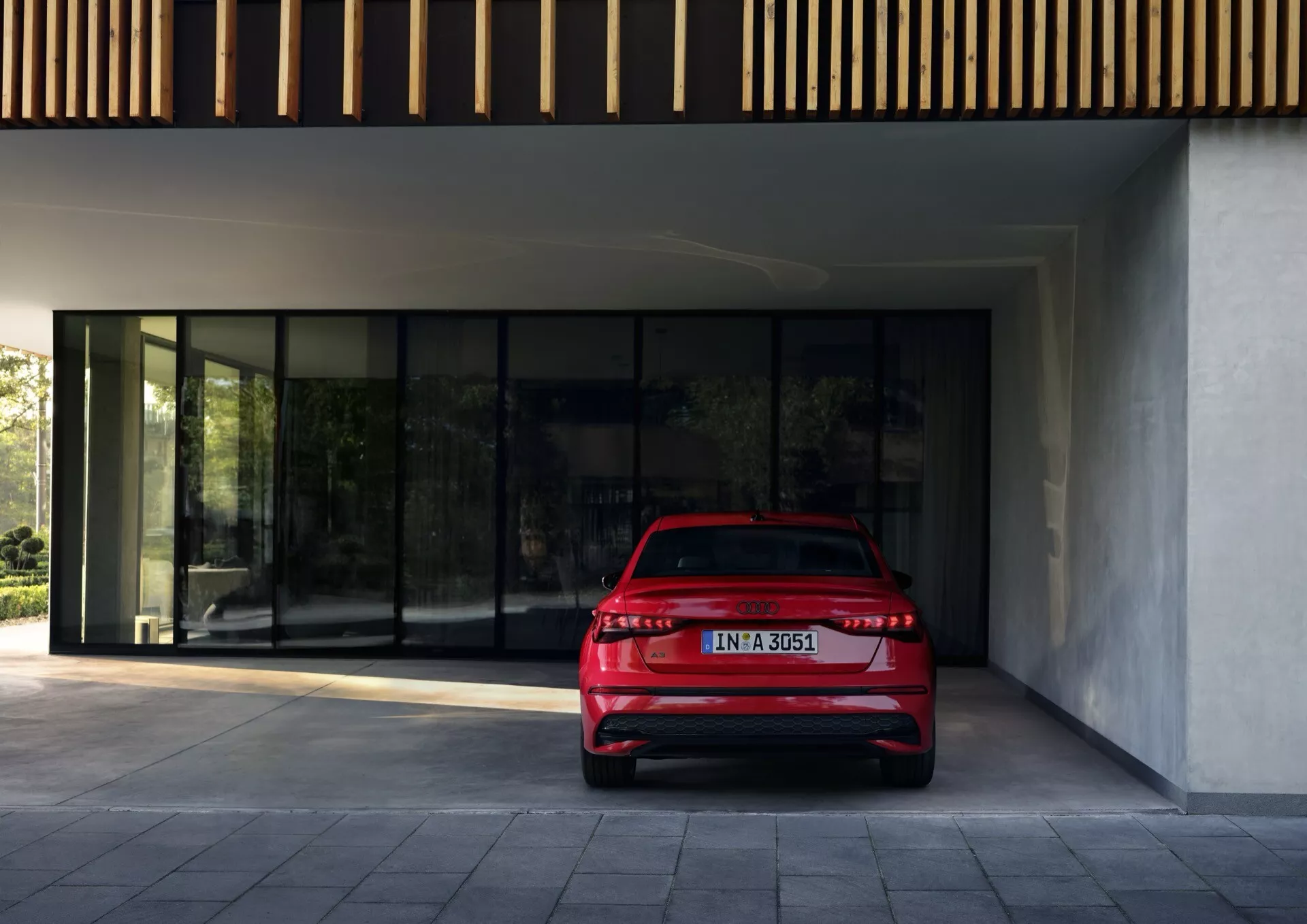 Červený Audi A3 (8Y-Facelift) 2024, fotografován z úhlu tři čtvrtě zadní části s důrazem na LED zadní světla, dvojitý výfukový systém a decentní zadní spoiler, zaparkovaný venku před moderní budovou