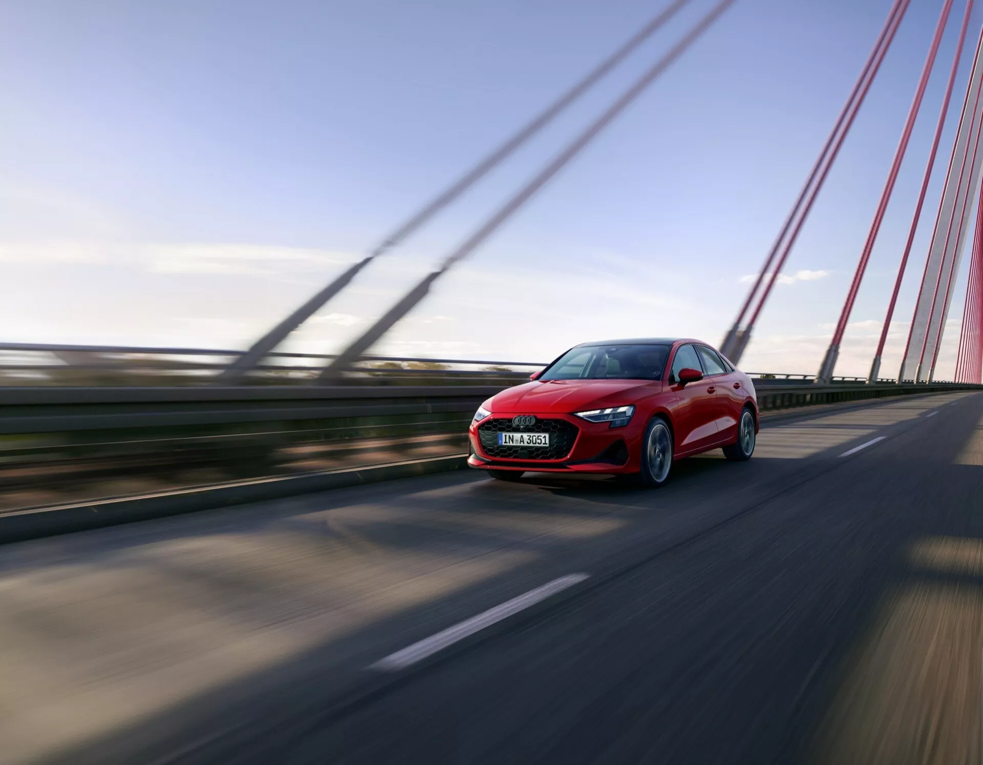 Audi A3 (8Y-Facelift) 2024 v pohybu na mostě, ukazující dynamický pohled zepředu a z boku pod nízkým úhlem pro dramatický efekt, zvýrazněný rozmazáním pohybu okolo rychle se pohybujícího vozidla, s viditelnými designovými prvkami jako jsou LED světlomety a aerodynamické kontury na pozadí jasné oblohy.