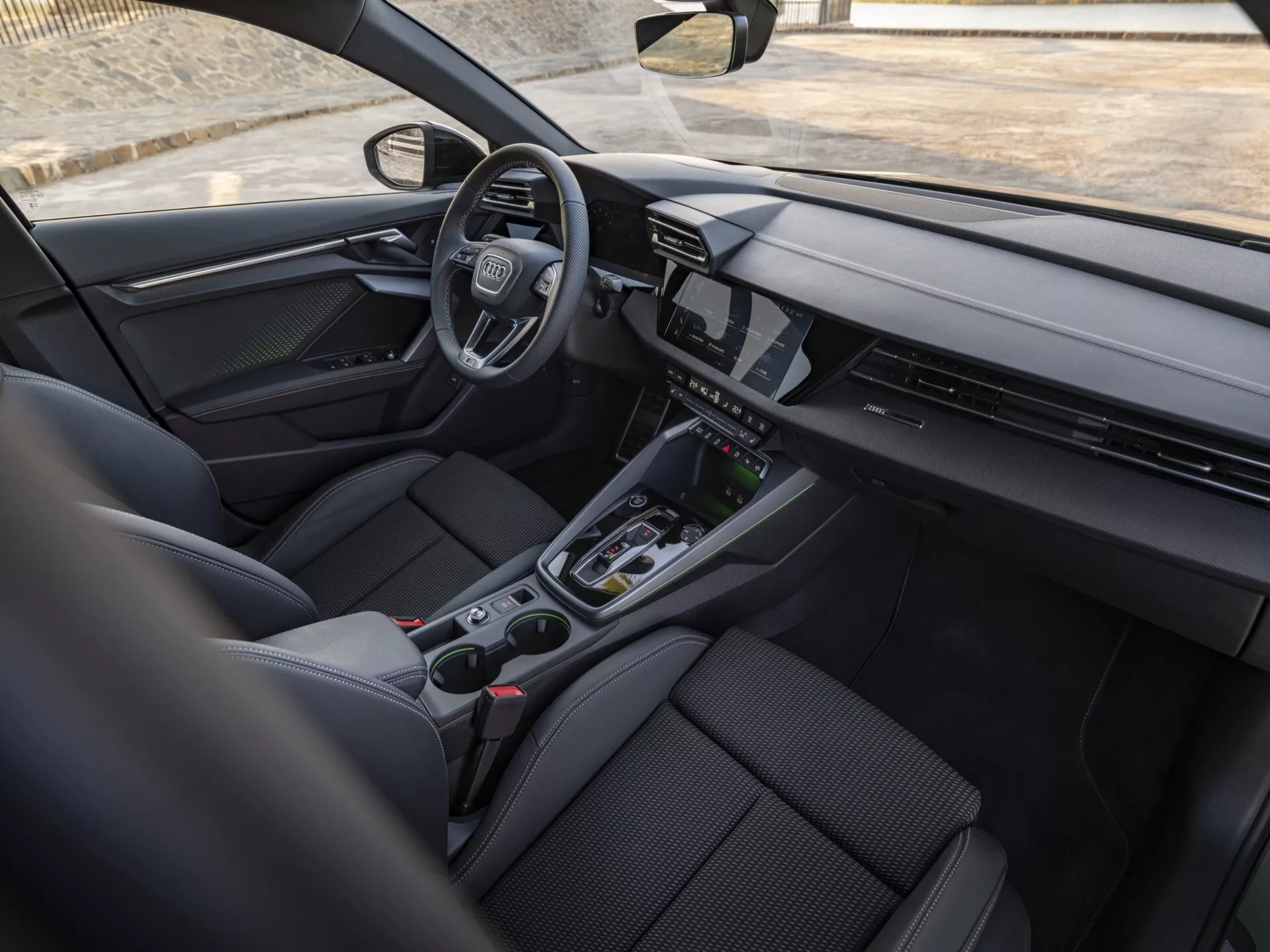 Interiér Audi A3 (8Y-Facelift) z roku 2024 z pohledu zadního pasažéra s elegantní přístrojovou deskou s nepřetržitým digitálním displejem, volantem na levé straně s emblémem, minimalistickými ovladači na středové konzole, sportovně ergonomickými předními sedadly a pokročilými technologickými funkcemi