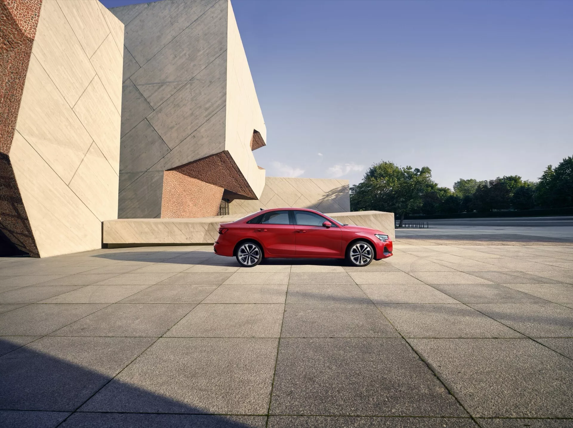 Facelift Audi A3 8Y 2024 ve světlé červené barvě, zobrazený z čelně-bočního úhlu, který zdůrazňuje elegantní sestavu LED světlometů, lesklou přední mřížku, aerodynamicky navržená vícepaprsková hliníková kola a moderní siluetu na pozadí městské kulisy s výraznými architektonickými prvky, vše zachycené v přirozeném slunečním světle