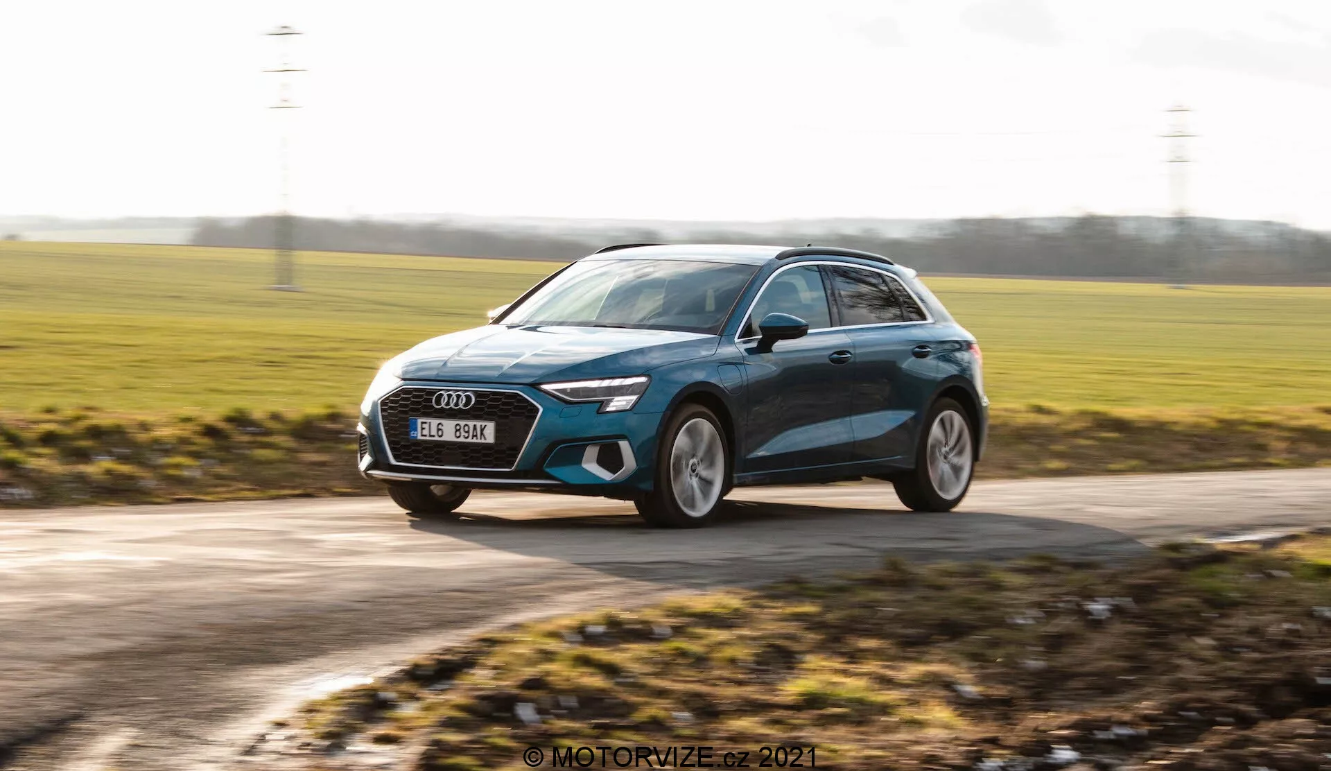 Dynamický záběr moderního hatchbacku, pravděpodobně Audi A3 Sportback z roku 2020, zachycený v pohybu. Obraz je pořízen z mírně nízkého úhlu, zdůrazňující výšku vozu a vytvářející dynamický pocit. Ukazuje tříčtvrteční přední pohled s vozem pohybujícím se zprava doleva. Mezi designové prvky patří elegantní LED světlomety, velká přední maska, velká litá kola, aerodynamická zrcátka s potenciálními blinkry a metalická barva s konturami. Pozadí je rozmazané, což zvyšuje pocit rychlosti, zatímco samotný vůz je v ostrém zaostření, což zvýrazňuje jeho moderní design a prémiovou estetiku.