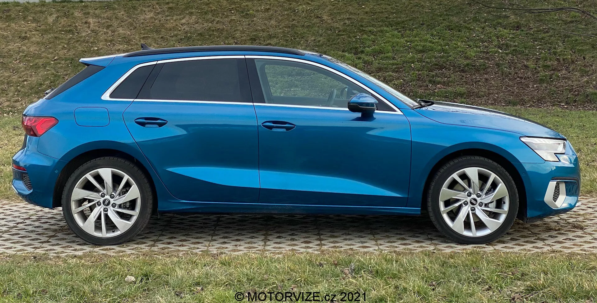 Boční profil modrého Audi A3 Sportback 2020, předvádějícího moderní hatchback design se sportovní klesající linií střechy, slitými koly, zavřenými okny a bočními zrcátky v barvě karoserie s integrovanými směrovkami, zaparkovaného na trávníku vedle chodníku.