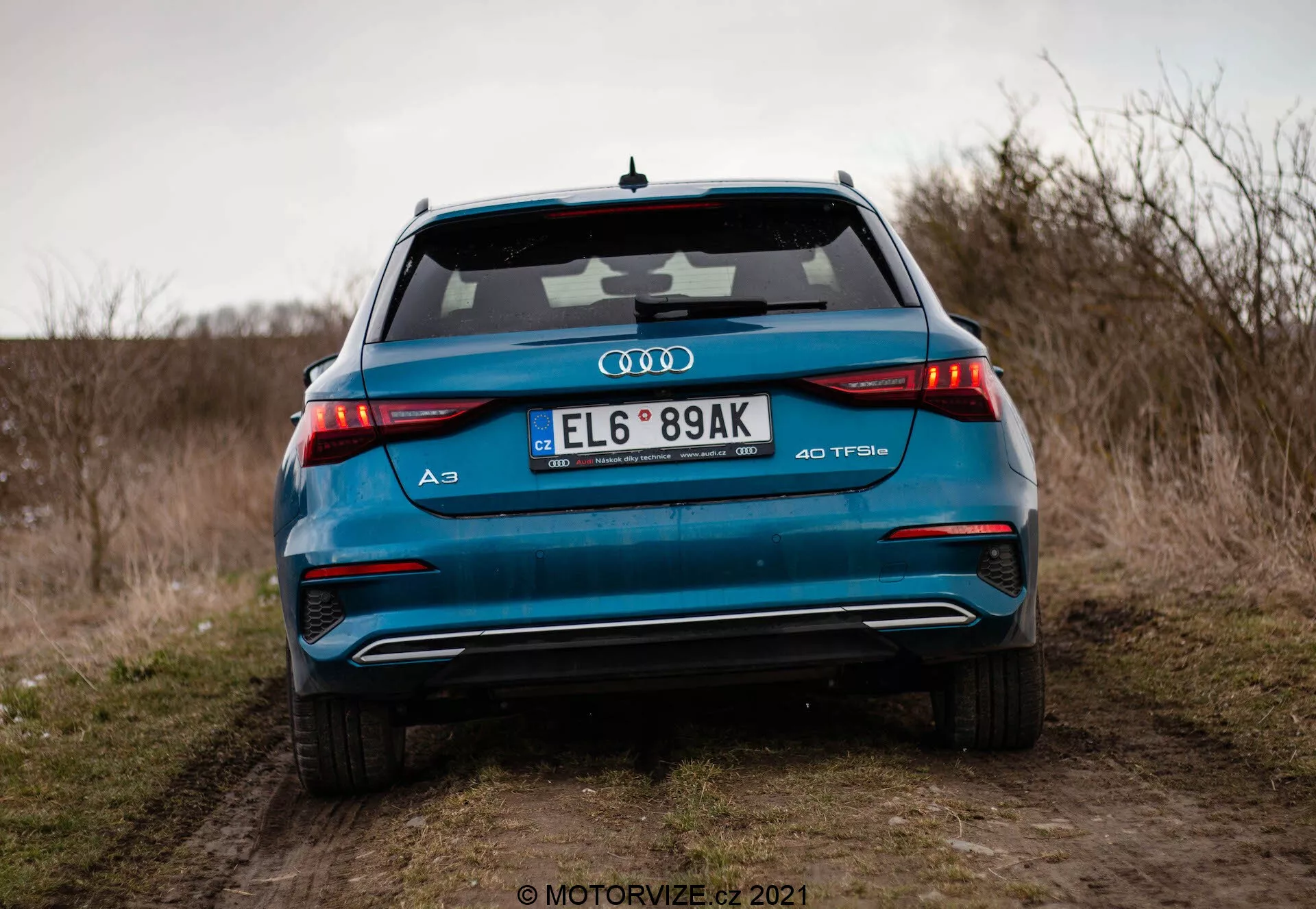 Detailní pohled na zadní část zářivě modrého Audi A3 Sportback 40 TFSI z roku 2020, zdůrazňující jeho pokročilé designové prvky včetně horizontálních LED zadních světel, dvojitého výfuku, znaku TFSI s turbomotorem, integrovaného spoileru a žraločí antény, fotografovaný z nízkého úhlu na prašné cestě s otevřeným polem a zataženou oblohou v pozadí.