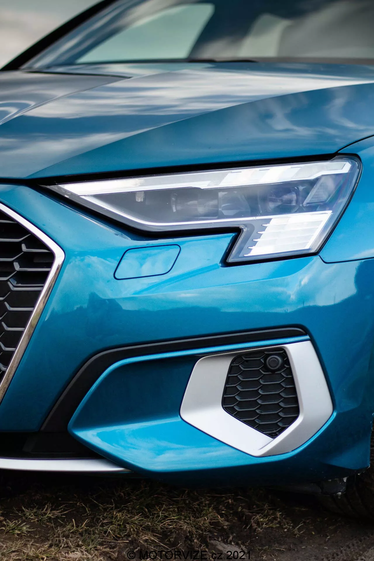 Detailní, nízkoúhlý záběr přední levé části Audi A3 Sportback z roku 2020, zachycující ostrý LED světlomet, šestiúhelníkovou mřížku s voštinovým vzorem, která může obsahovat senzory nebo odznaky, a část lesklé jasně modré kapoty a nárazníku, zdůrazňující moderní, sofistikovaný design a pokročilou automobilovou technologii.