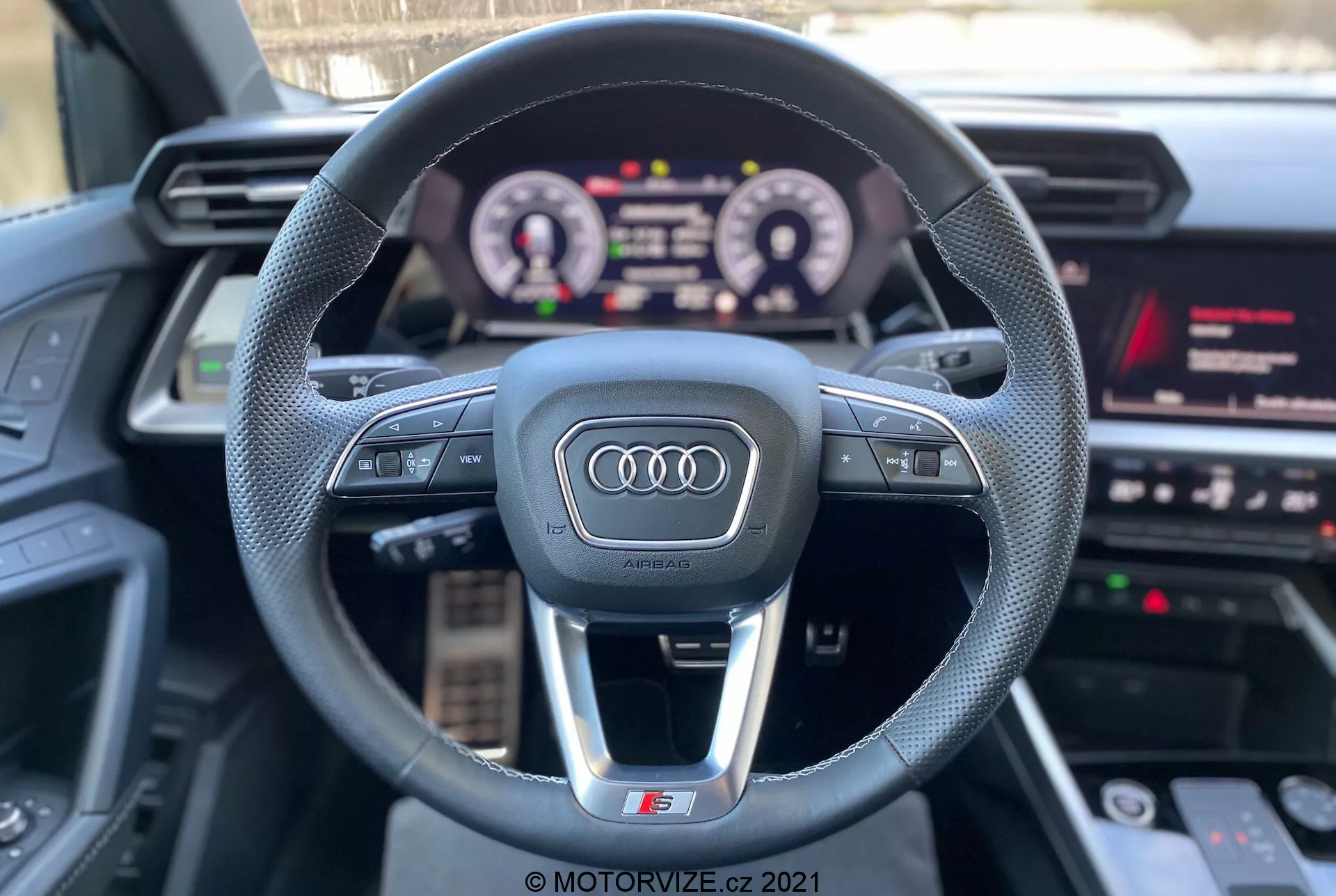 Pohled z první osoby ze sedadla řidiče vozu Audi A3 2020 (A3 Sportback), který ukazuje sportovní tříramenný volant s logem Audi, multifunkčními ovládacími tlačítky, osvětlenou digitální přístrojovou deskou a částí infotainment systému s médii, navigací a nastavením vozidla. Vlastnosti zdůrazňují interaktivní technologii a luxusní komponenty s důrazem na zážitek z jízdy.