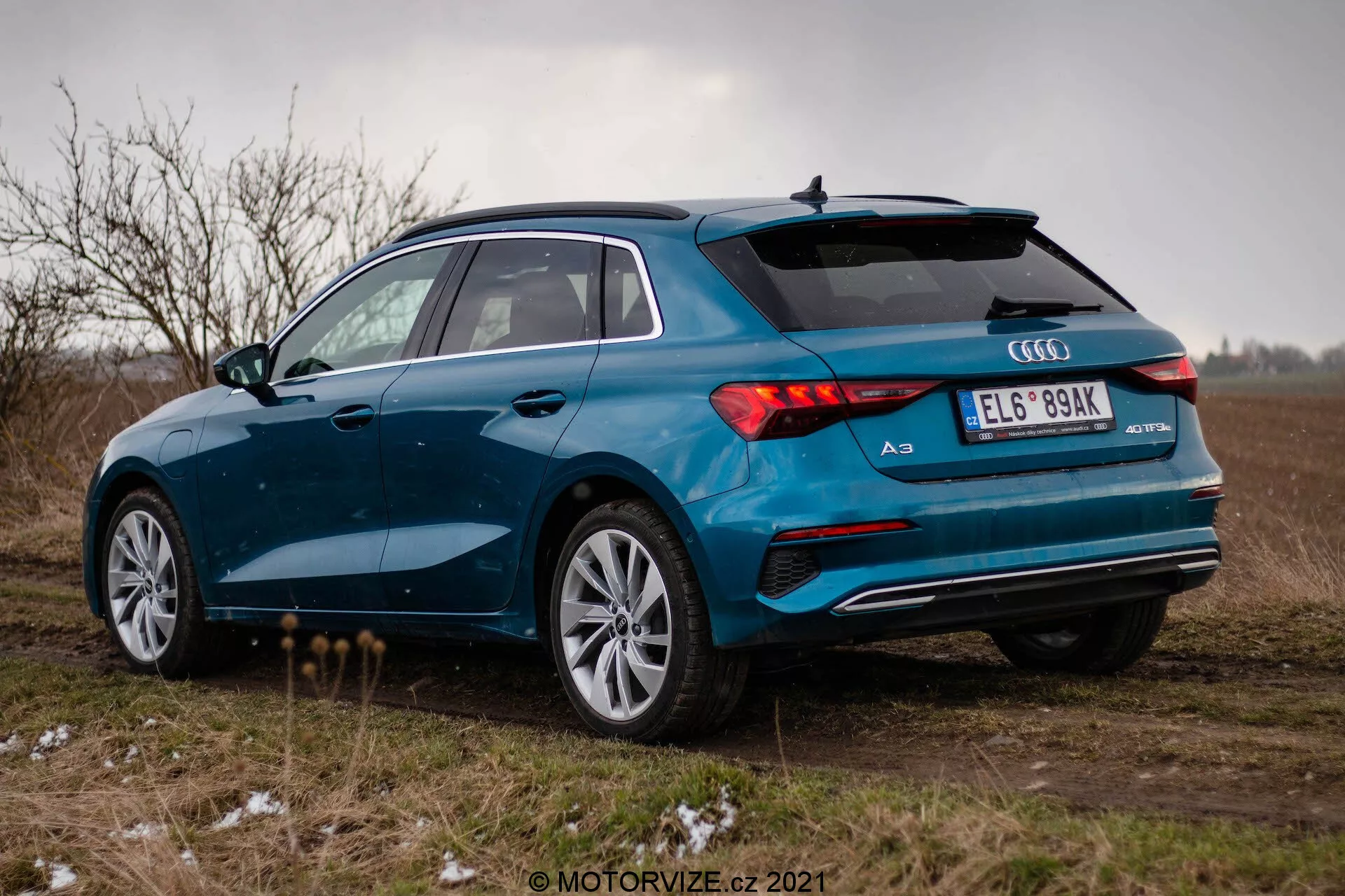 Modré Audi A3 2020 (Sportback) fotografované ze tříčtvrtinového úhlu zezadu na otevřeném poli pod zamračenou oblohou. Obrázek zvýrazňuje pravou stranu vozidla, zadní LED světla, dvojitý výfukový systém, vícepaprsková hliníková kola a zadní spoiler. Vozidlo je zaparkováno na neasfaltovém povrchu s trsy trávy a lehkou vrstvou sněhu nebo námrazy, což naznačuje chladné počasí. Celková nálada je ponurá s důrazem na sportovní a prémiový design Audi A3.