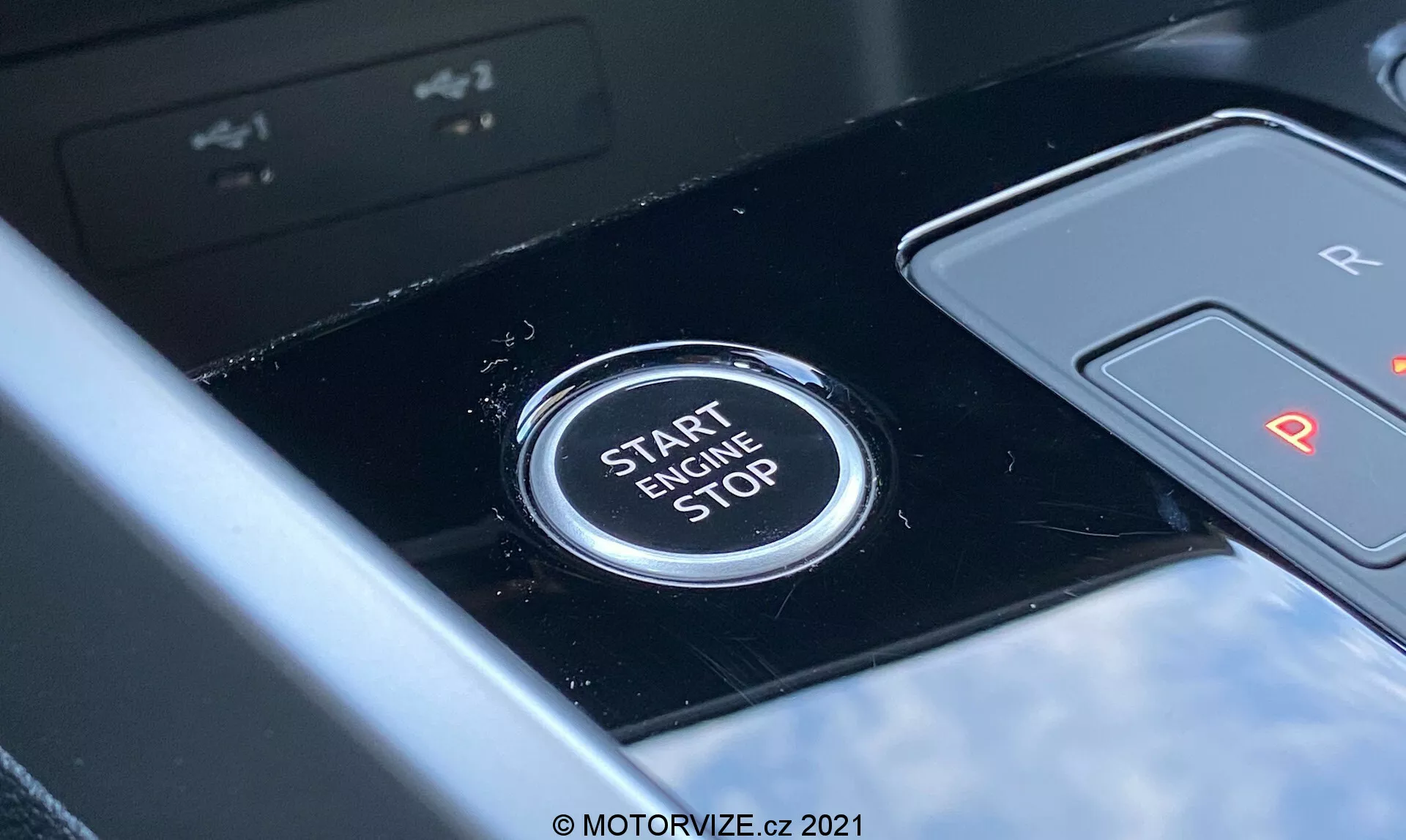 Detailní pohled na tlačítko start/stop motoru Audi A3 Sportback 2020 umístěné ve středové konzole. Tlačítko zřetelně zobrazuje text 'START ENGINE STOP' velkými písmeny, což naznačuje jeho funkci pro spuštění nebo vypnutí motoru. Pohled z perspektivy řidiče, lesklý černý panel a částečně viditelná řadicí páka s označením 'R' a 'P' naznačují automatickou převodovku. Mělká hloubka ostrosti zdůrazňuje jasnost tlačítka oproti lehce rozmazanému pozadí, což poukazuje na moderní interiér vozidla s pokročilými technologickými funkcemi.
