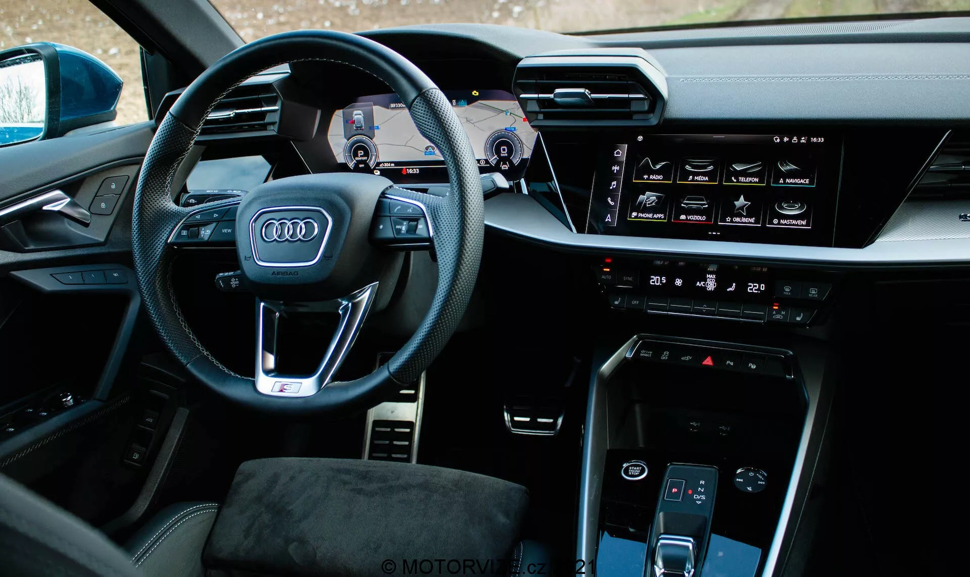 Pohled do interiéru Audi A3 Sportback 2020 z mírně posunutého levého úhlu zaměřený na stranu řidiče. Prominentní volant nese ikonické logo čtyř kruhů Audi, multifunkční tlačítka a viditelné páčky řazení. Za volantem se nachází digitální přístrojový panel nazývaný Audi Virtual Cockpit, který zobrazuje tachometr, otáčkoměr a navigační mapu. Středová konzola obsahuje horizontální dotykový infotainment display, ovládání klimatizace, tlačítko start-stop motoru, další dotykový displej pro ovládání klimatizace a minimalistický volič převodovky. Interiér se vyznačuje vysoce kvalitními materiály a technologicky integrovaným designem s prémiovým vzhledem.