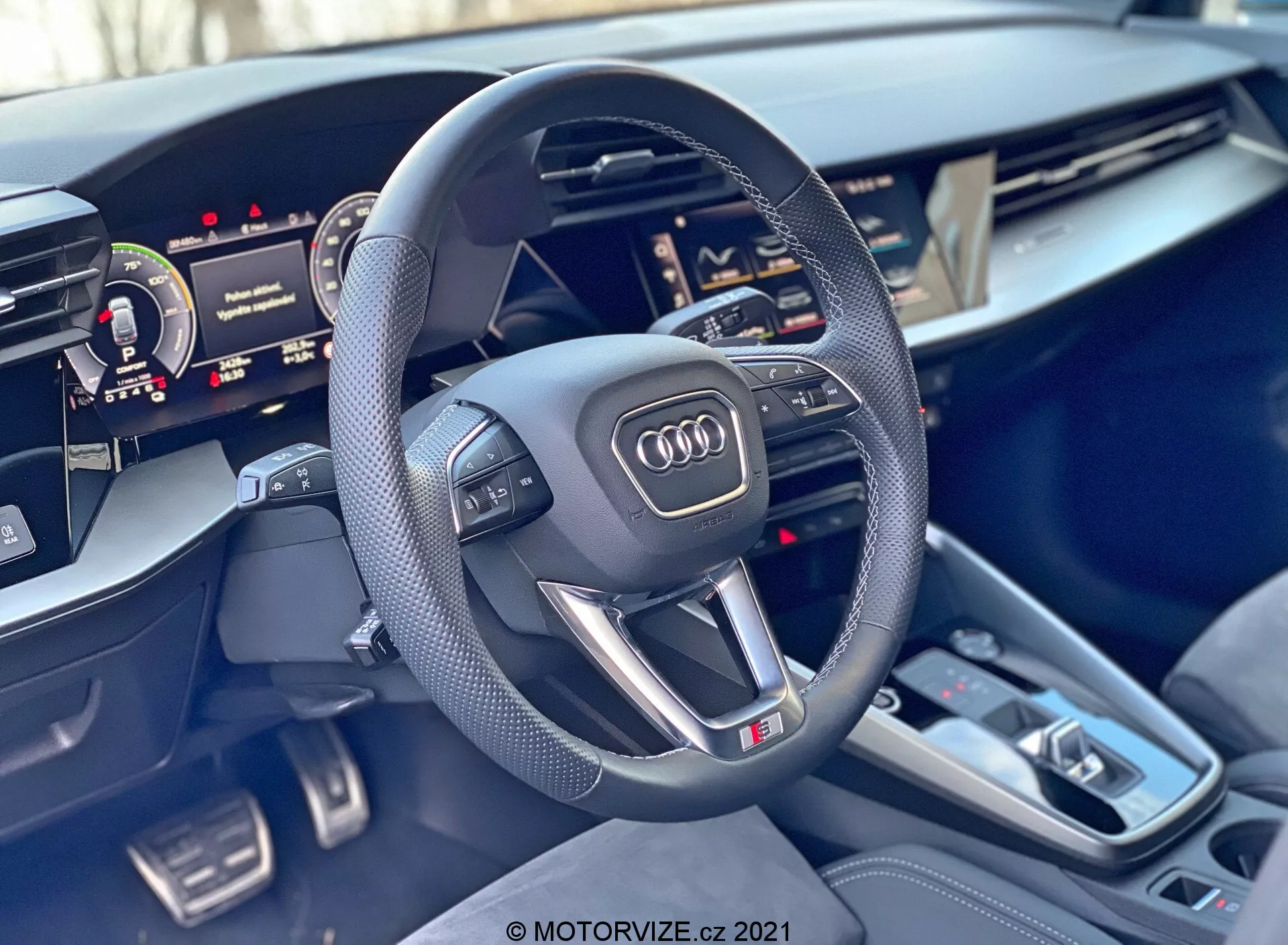 Pohled do interiéru Audi A3 Sportback 2020 se zaměřením na volant a středovou konzoli. Sportovní tříramenný volant se sploštělou spodní částí má perforované kožené úchyty, multifunkční tlačítka, emblém Audi a značku 'S'. Za volantem je částečně viditelný digitální přístrojový panel a na pravé straně částečně zobrazený dotykový displej Centrálního multimediálního rozhraní (MMI). Středová konzola obsahuje řadicí páku automatické převodovky, několik tlačítek, dotykovou plochu, dva držáky nápojů a malé úložné přihrádky. Interiérové materiály mají prémiové nádechy kartáčovaného kovu a kvalitní provedení, zdůrazňující luxus a pokročilé technologie vozu.