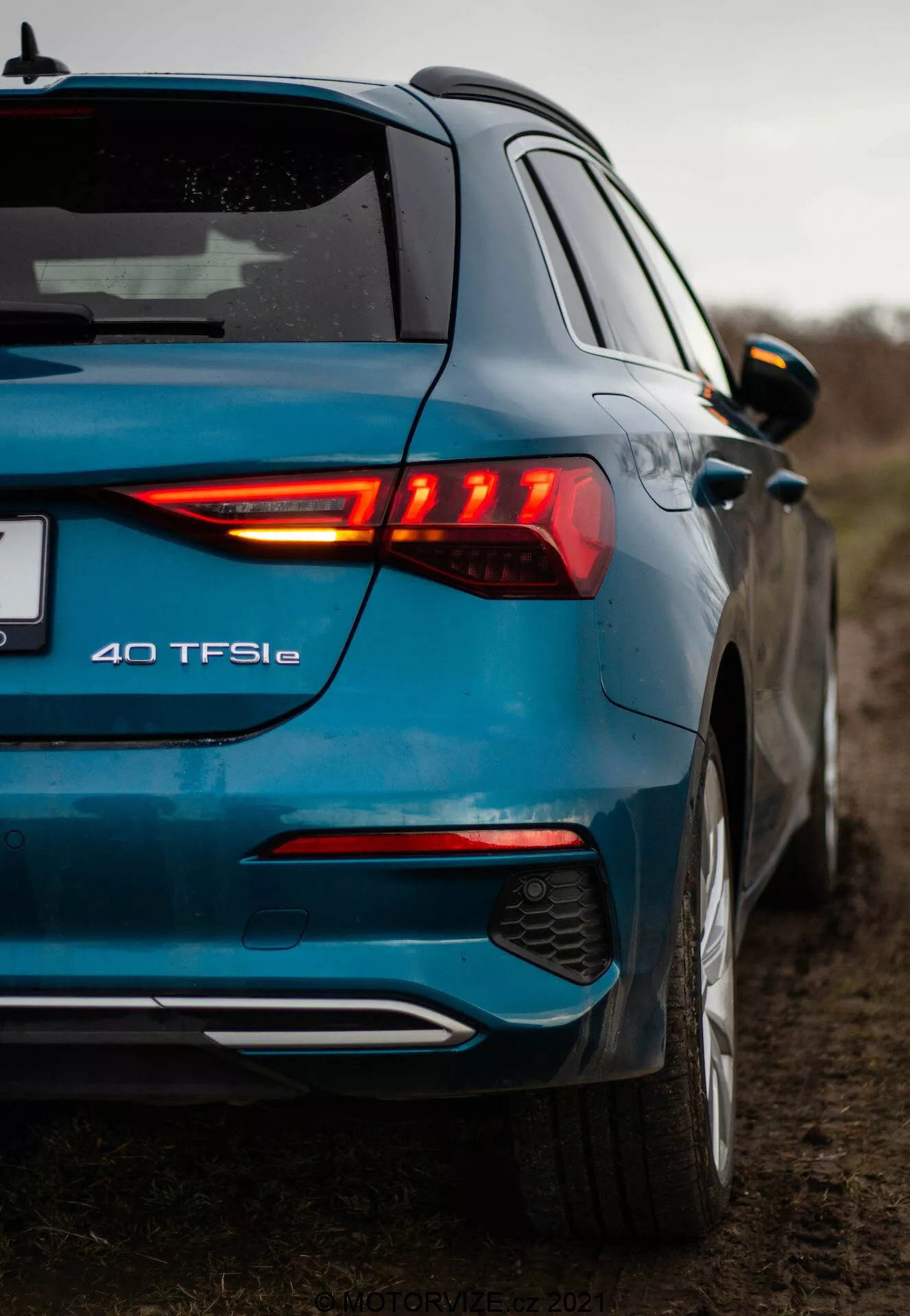 Nízký úhel pohledu na moderní modrý Audi A3 2020 (A3 Sportback) focený ze zadního tříčtvrtečního pohledu, zdůrazňující osvětlený zadní LED světelný klastr, odznak modelu 40 TFSIe, dvoutónový zadní nárazník s integrovaným difuzérem a parkovacími senzory, metalický lak, střešní spoiler, barevně sladěná boční zrcátka s integrovanými směrovkami a náznak předních dveří a bočního profilu. Fotografie zdůrazňuje designové prvky a značení vozidla v venkovském prostředí s zataženým nebem a malou hloubkou ostrosti.
