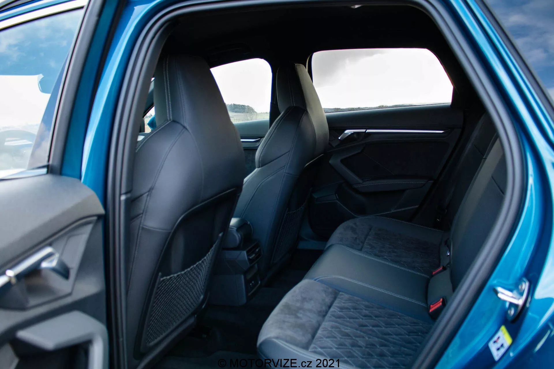 Záběr interiéru Audi A3 Sportback 2020 z pohledu zadních dveří ukazuje prostorné sedadla pro tři osoby s opěrkami hlavy a bezpečnostními pásy, čalouněné tmavou látkou se vzorem. Částečně viditelné jsou i přední sedadla s ergonomickým designem. Viditelné prvky dveřního panelu zahrnují rukojeť a možná ovládací prvky. Dobře osvětlený snímek zvýrazňuje elegantní, prémiový pocit s kvalitními materiály a designovými detaily.