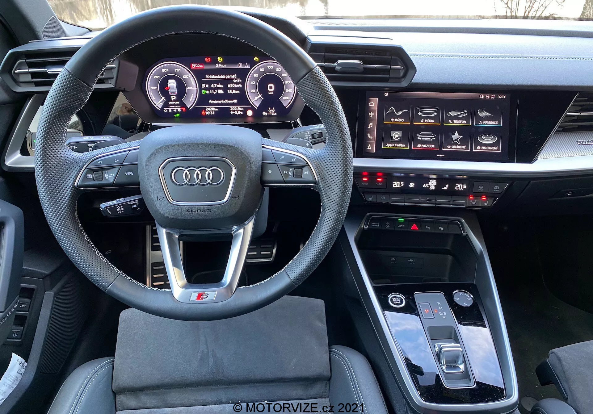 Pohled ze sedadla řidiče na interiér Audi A3 Sportback 2020 zobrazující sportovní multifunkční volant obložený kůží s plochým spodkem, digitální přístrojovou desku Audi Virtual Cockpit, významný infotainment systém, hmatová tlačítka pro klimatizaci a dotykovou podložku MMI v středové konzole, naznačující moderní, luxusní a technologicky vyspělé interiéry automobilů.