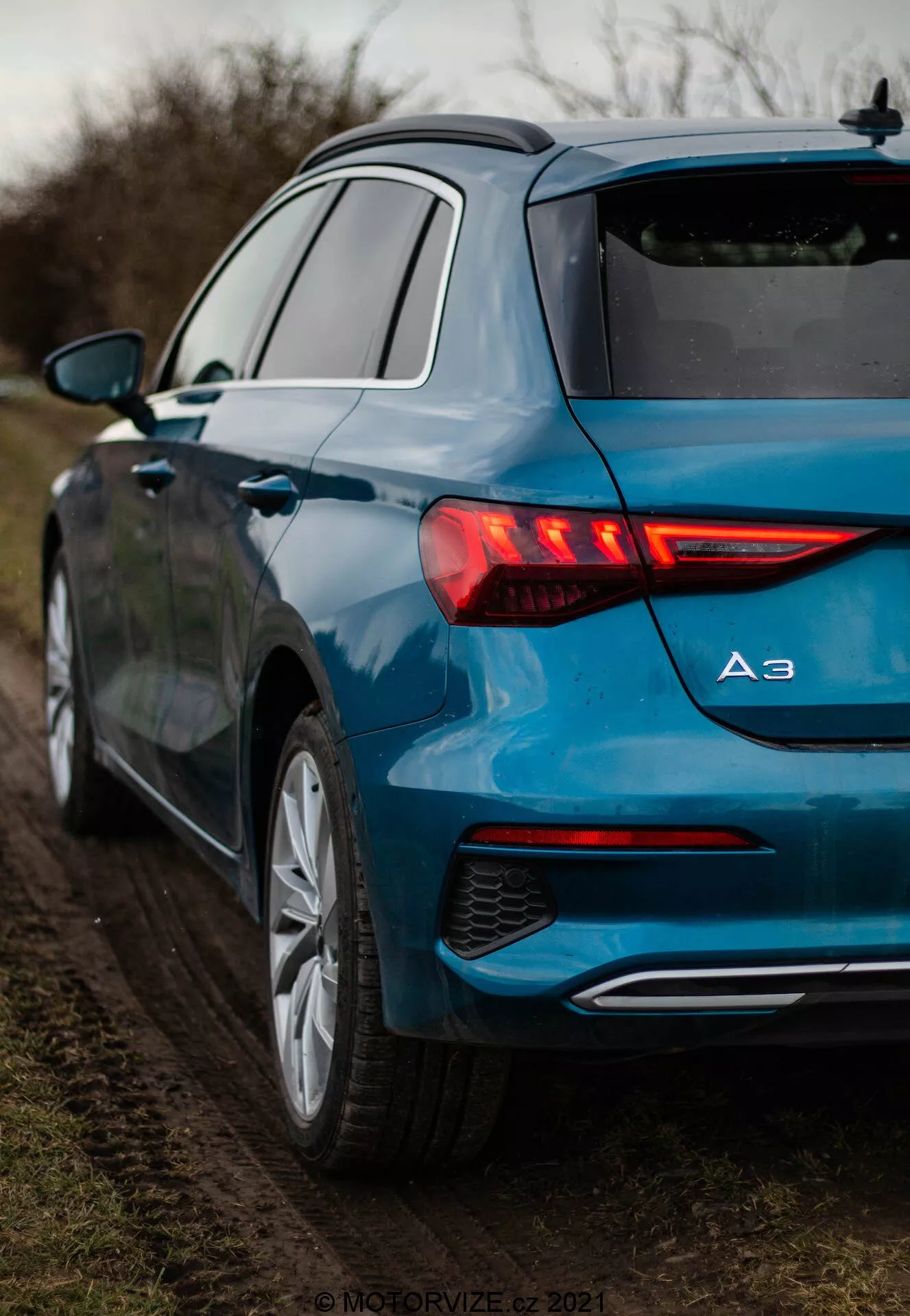 Detailní pohled na metalickou modrou Audi A3 2020 Sportback ze zadního tříčtvrtečního pohledu, zdůrazňující její osvětlená LED zadní světla s dynamickými směrovkami, modelový odznak zobrazující 'A3', vícepaprsková litá kola a jediný výfuk. Auto je zaparkované na nezpevněné krajnici obklopené vegetací, za měkkého, rozptýleného osvětlení.
