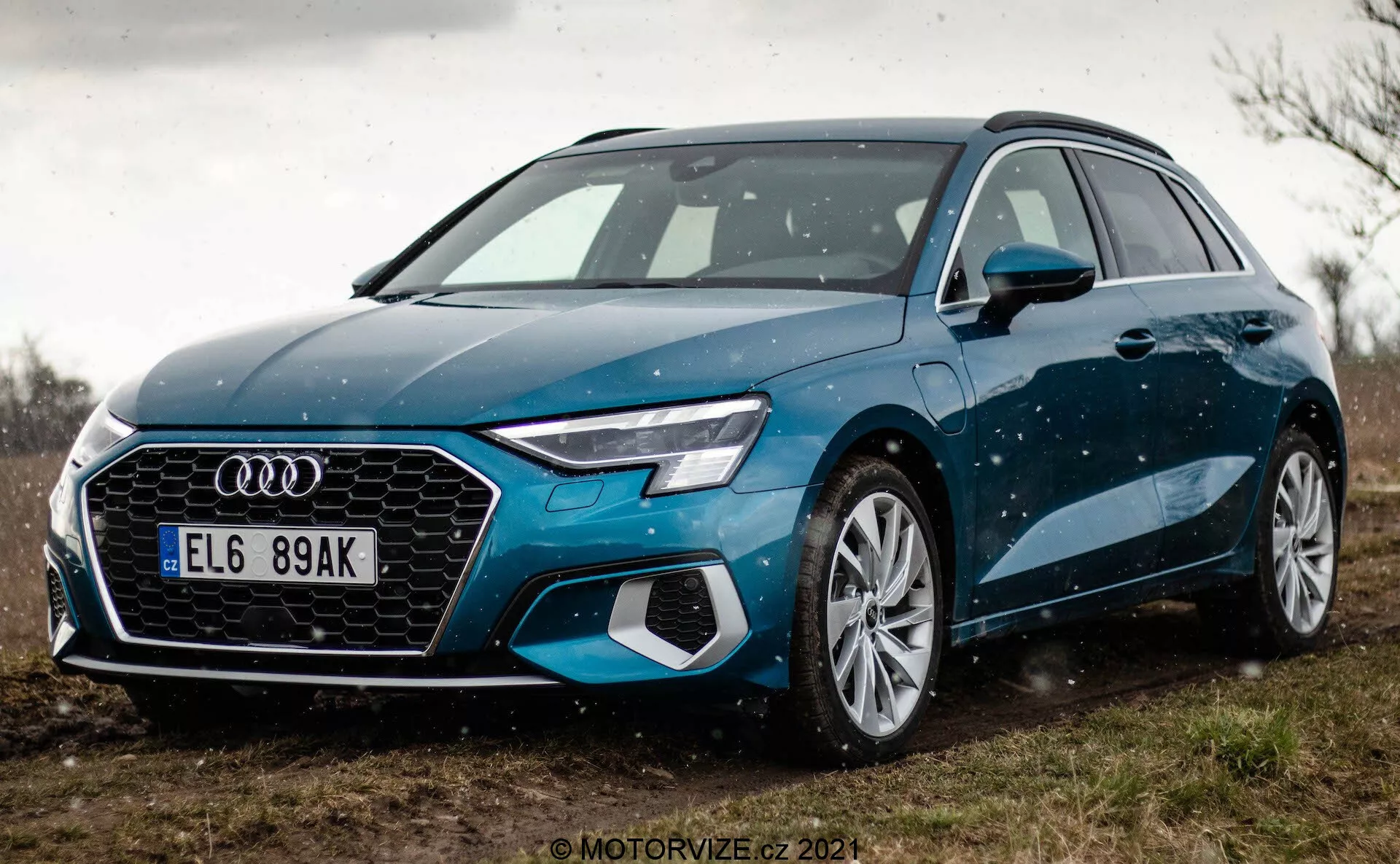 Pohled z nízkého úhlu na modrou Audi A3 Sportback 2020, zdůrazňující přední masku a design LED světlometů. Blízky záběr se zaměřuje na přední levou stranu vozu, ukazující jeho hexagonální masku s výrazným znakem Audi, integrovanými senzory pro funkce ADAS, velká kola z lehké slitiny s více paprsky a elegantní boční zrcátka. Auto je zaparkováno na nezpevněném povrchu s vegetací v pozadí, což naznačuje venkovské prostředí. Jemné kapky vody na karoserii vozu zvýrazňují jeho linie a barvu.
