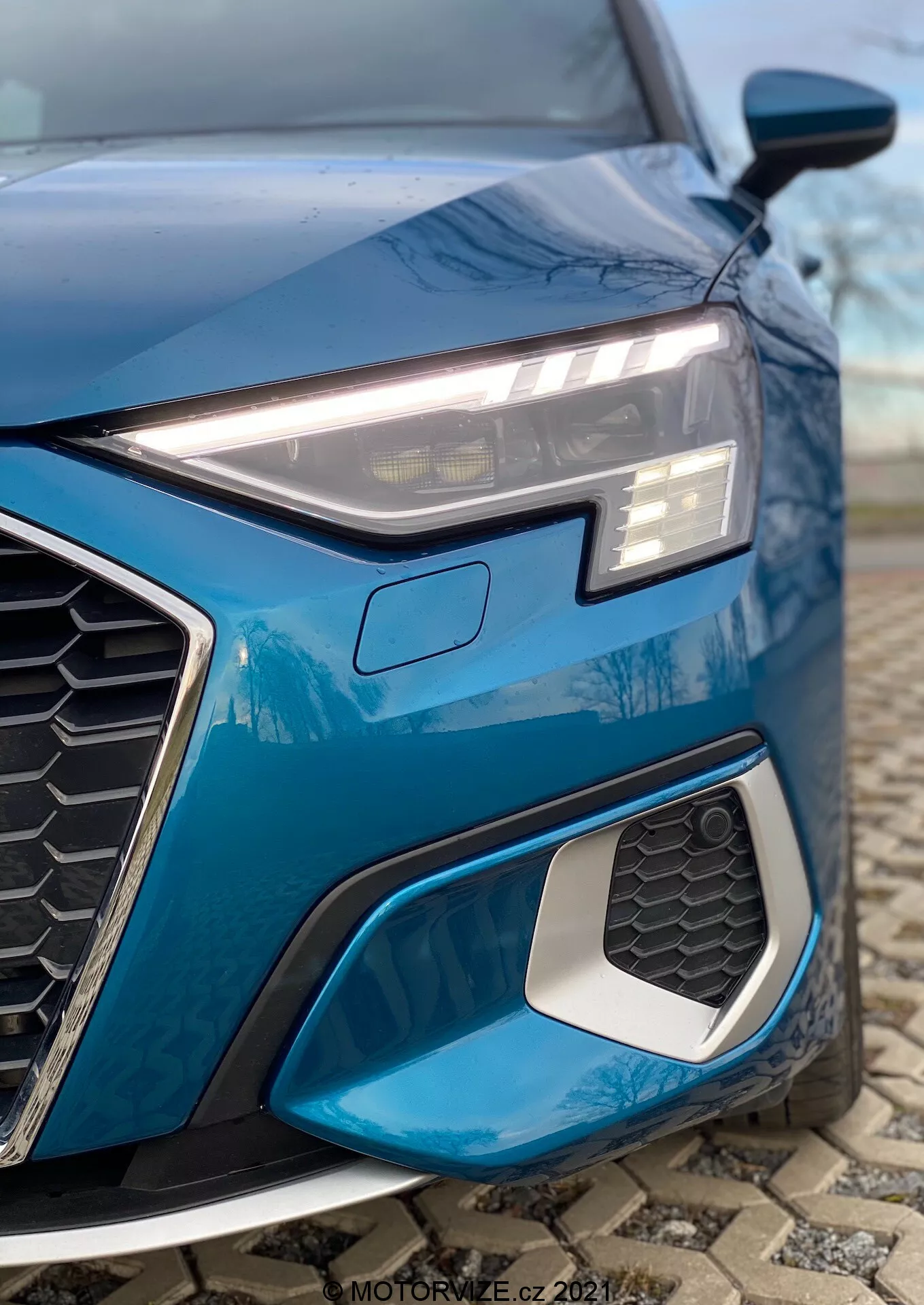 Detailní záběr na levý přední čtvrt auta modrého Audi A3 2020 (A3 Sportback) s důrazem na elegantní, úhlovou LED světlomety s denním svícením, výraznou hexagonální masku chladiče s motivem včelích plástů a sání vzduchu s kovovým lemem. Nízký úhel záběru zdůrazňuje sportovní designové prvky, složitý geometrický design mlhových světel a rozostřené pozadí pro zaměření na detaily.